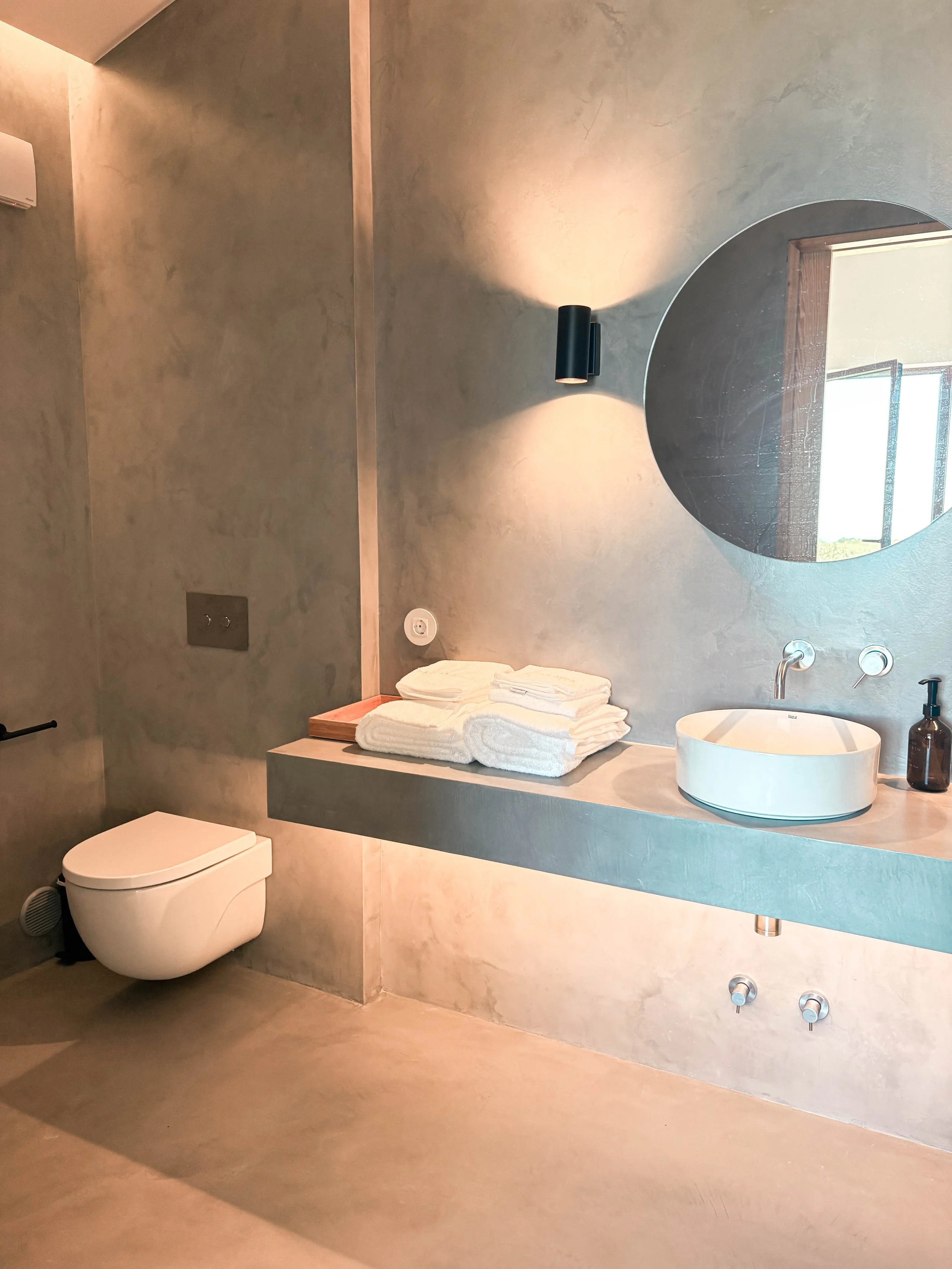 Banheiro com parede de tom bege, pia de balcão de concreto com cuba redonda branca, espelho redondo, toalhas brancas dobradas, torneira moderna, espelho grande na parede, luz de parede, vaso sanitário moderno, janela visível no espelho, dispenser de 