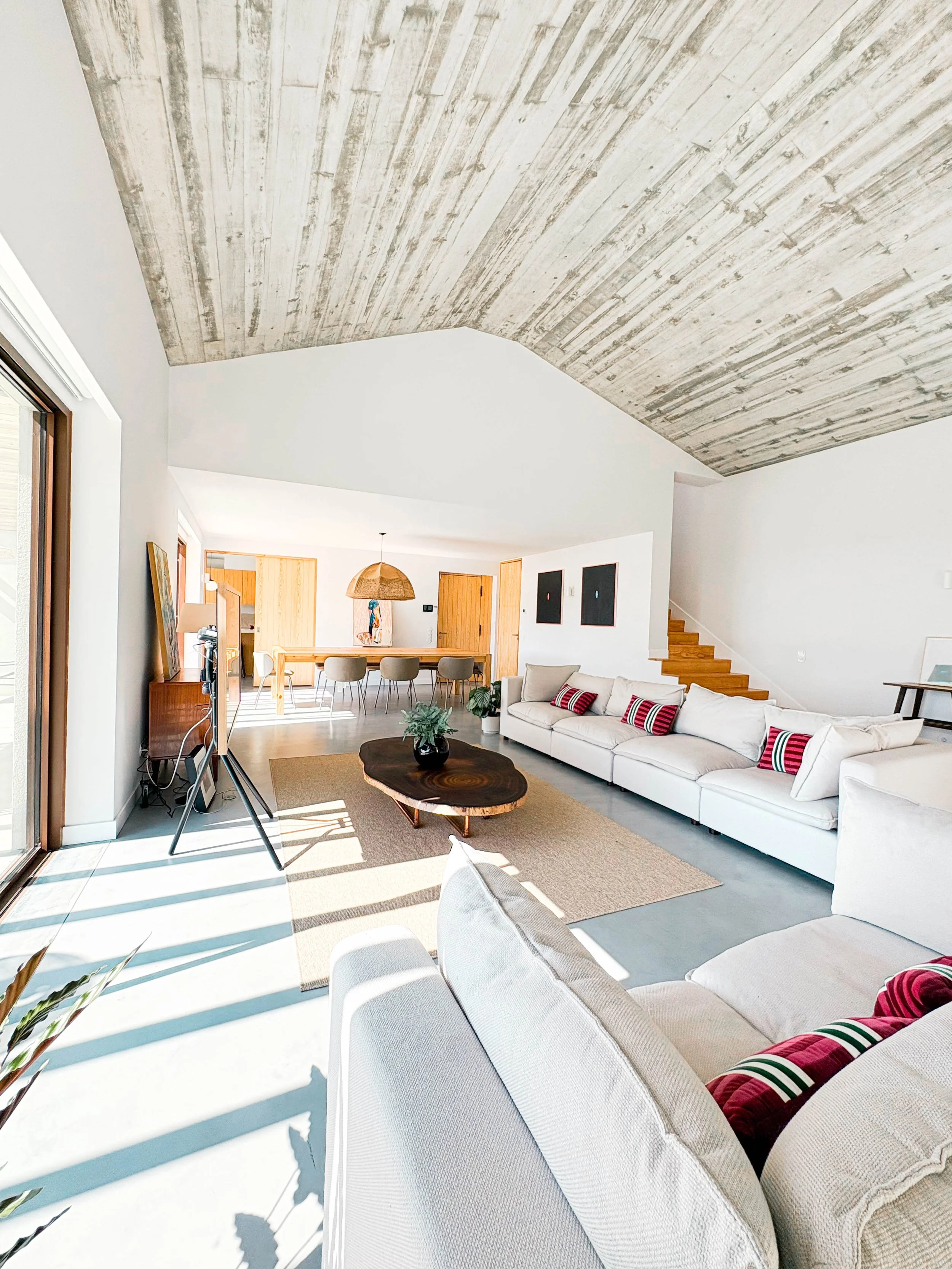 Sala de estar moderna com teto de concreto, sofá branco com almofadas vermelhas com listras, mesa de centro de madeira natural, janelas grandes com luz natural, área de jantar ao fundo com mesa de madeira e cadeiras, escada de madeira ao lado, decora