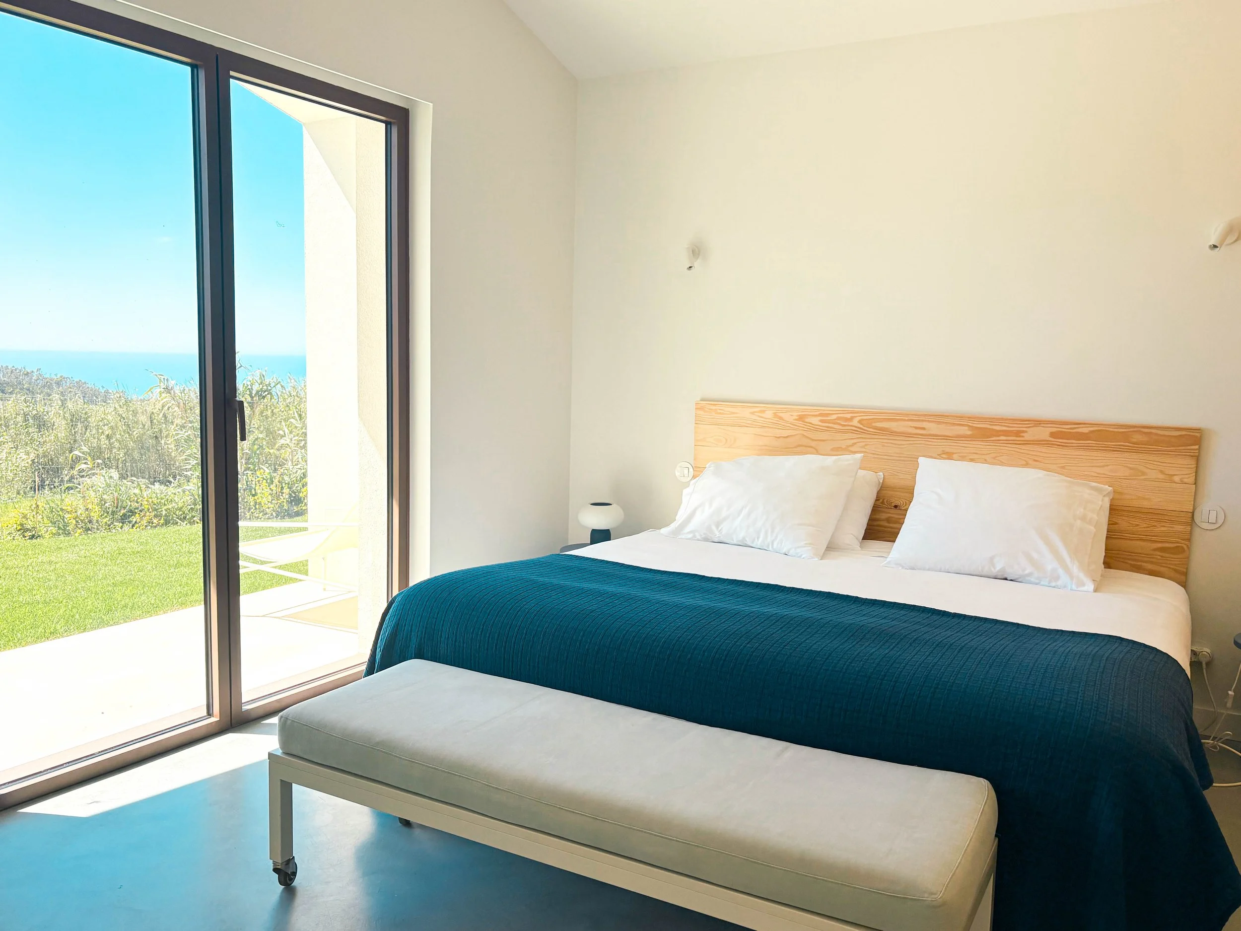 Quarto com cama de casal com cabeceira de madeira, roupa de cama branca, edredom azul escuro e duas almofadas brancas, ao lado de uma janela de vidro que revela uma vista de céu, mar e natureza, uma mesa de cabeceira com luminária ao lado da cama, e 