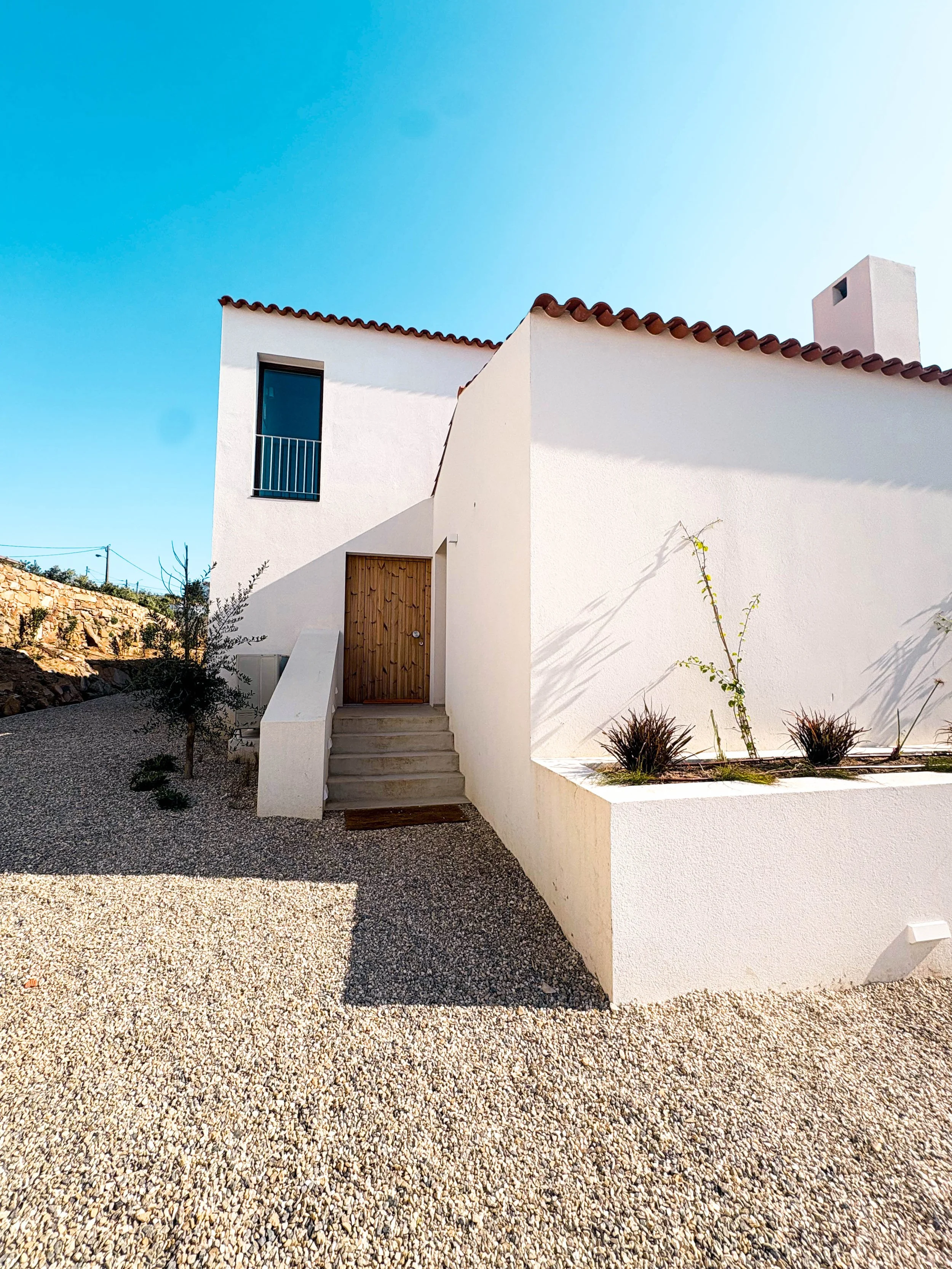 Imagem de uma casa moderna com paredes brancas, telhado de barro e uma escada de concreto na entrada, sob céu azul claro.