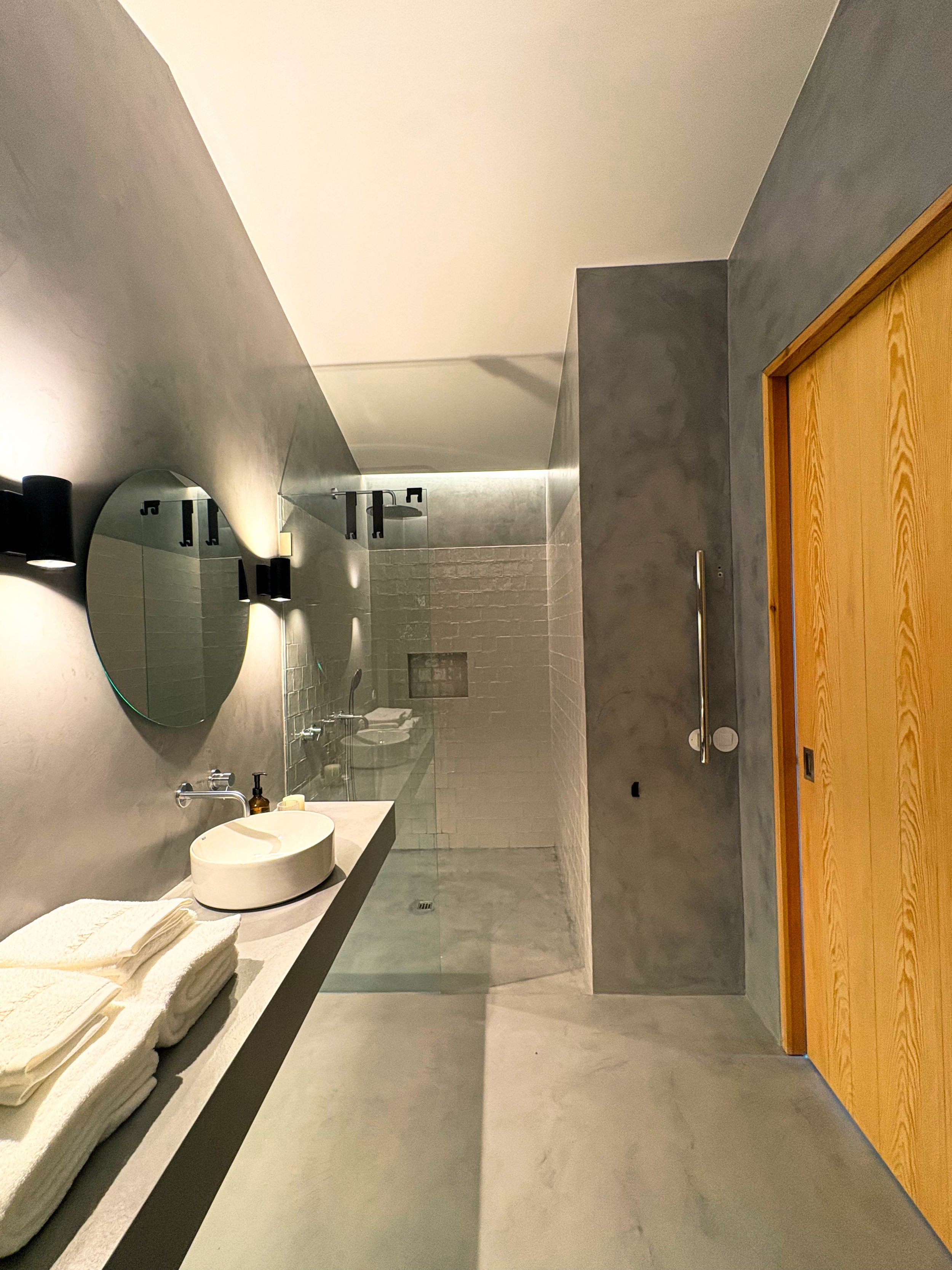 Banheiro moderno com parede de cimento, espelho redondo acima da pia, toalhas brancas dobradas, pia de porcelana branca, ducha de vidro e porta de madeira.