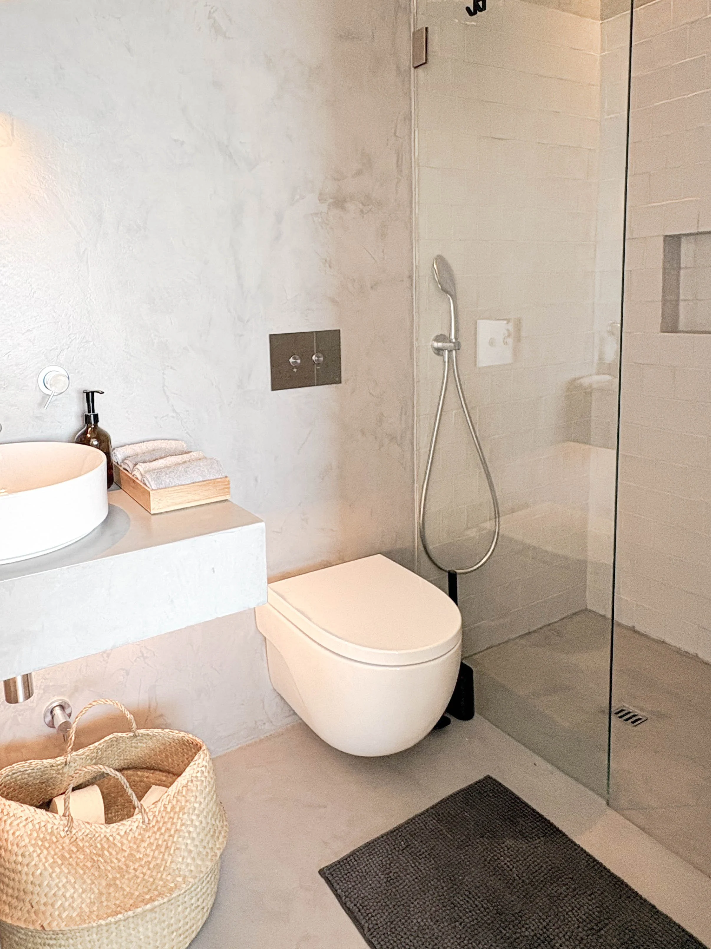 Banheiro com vaso sanitário, pia com bancada, cesta de limpeza, toalhas, ducha no box e espelho.