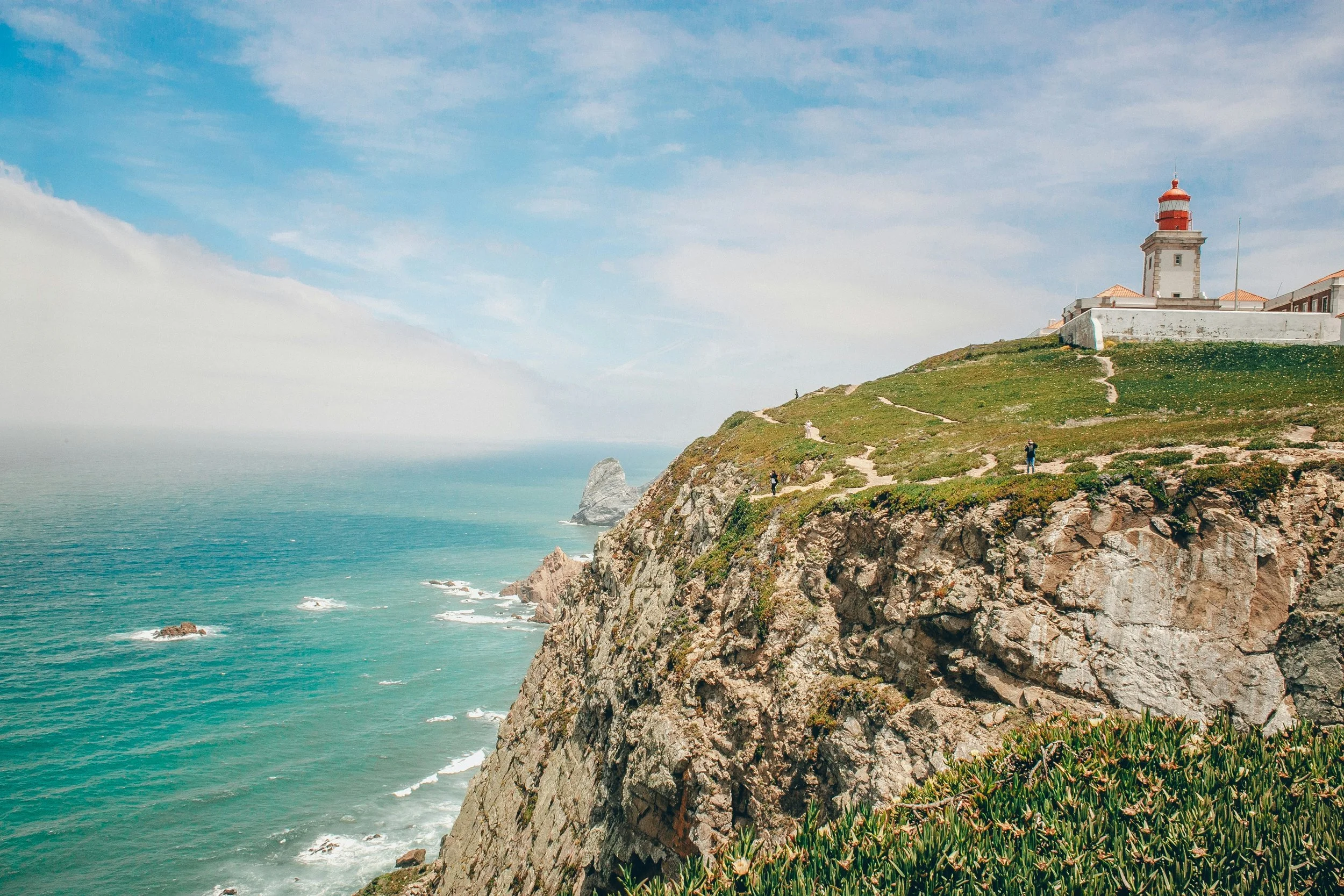 Faro em cima de um penhasco com vista para o oceano, céu azul com nuvens e vegetação ao redor.
