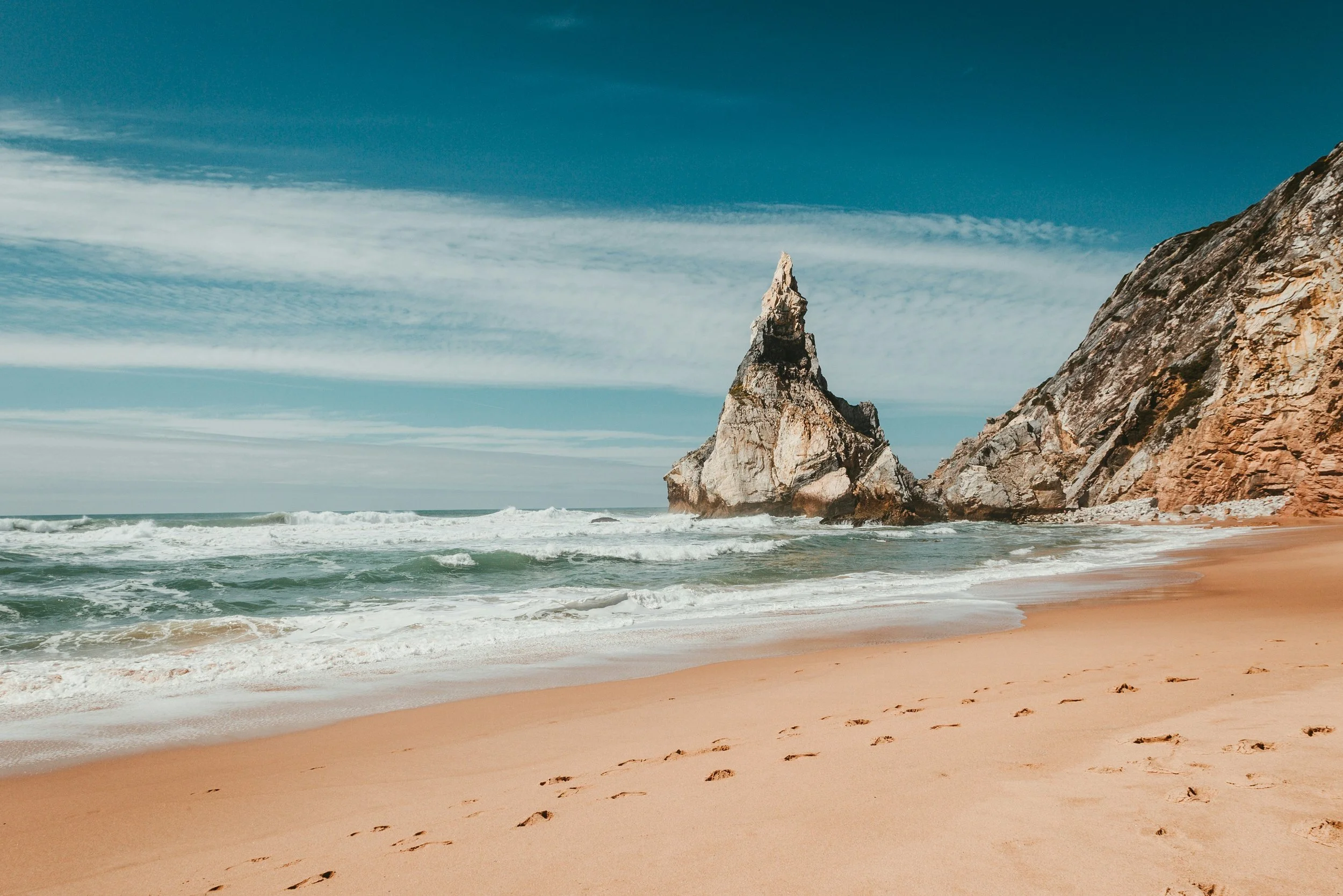 Praia com areia dourada, ondas do mar, rochas grandes e uma formação rochosa pontiaguda ao fundo, sob céu parcialmente nublado.