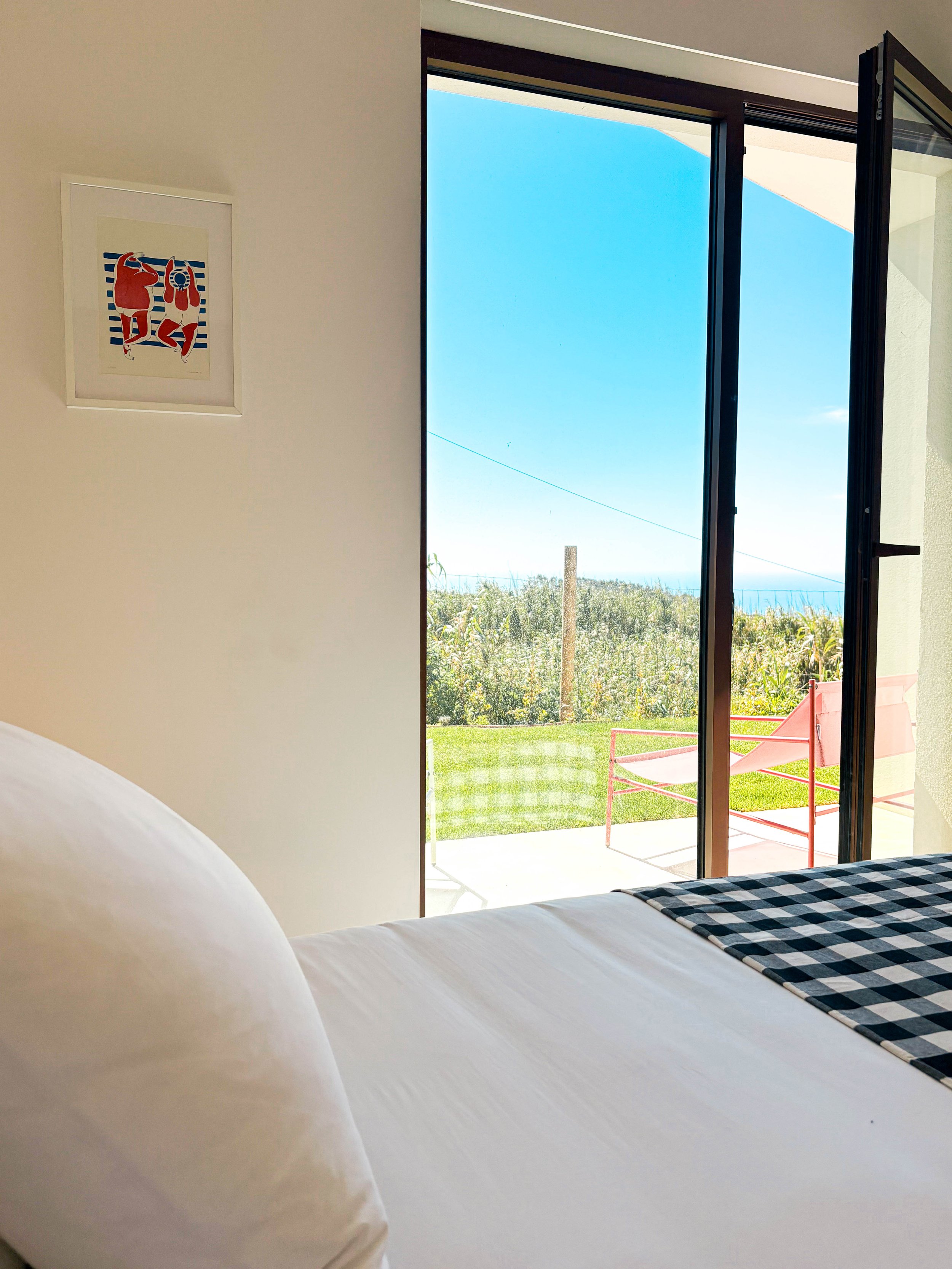 Quarto com cama com colcha de xadrez, parede branca, quadro com figuras humanas, porta de vidro aberta mostrando jardim, céu azul e mar ao fundo.