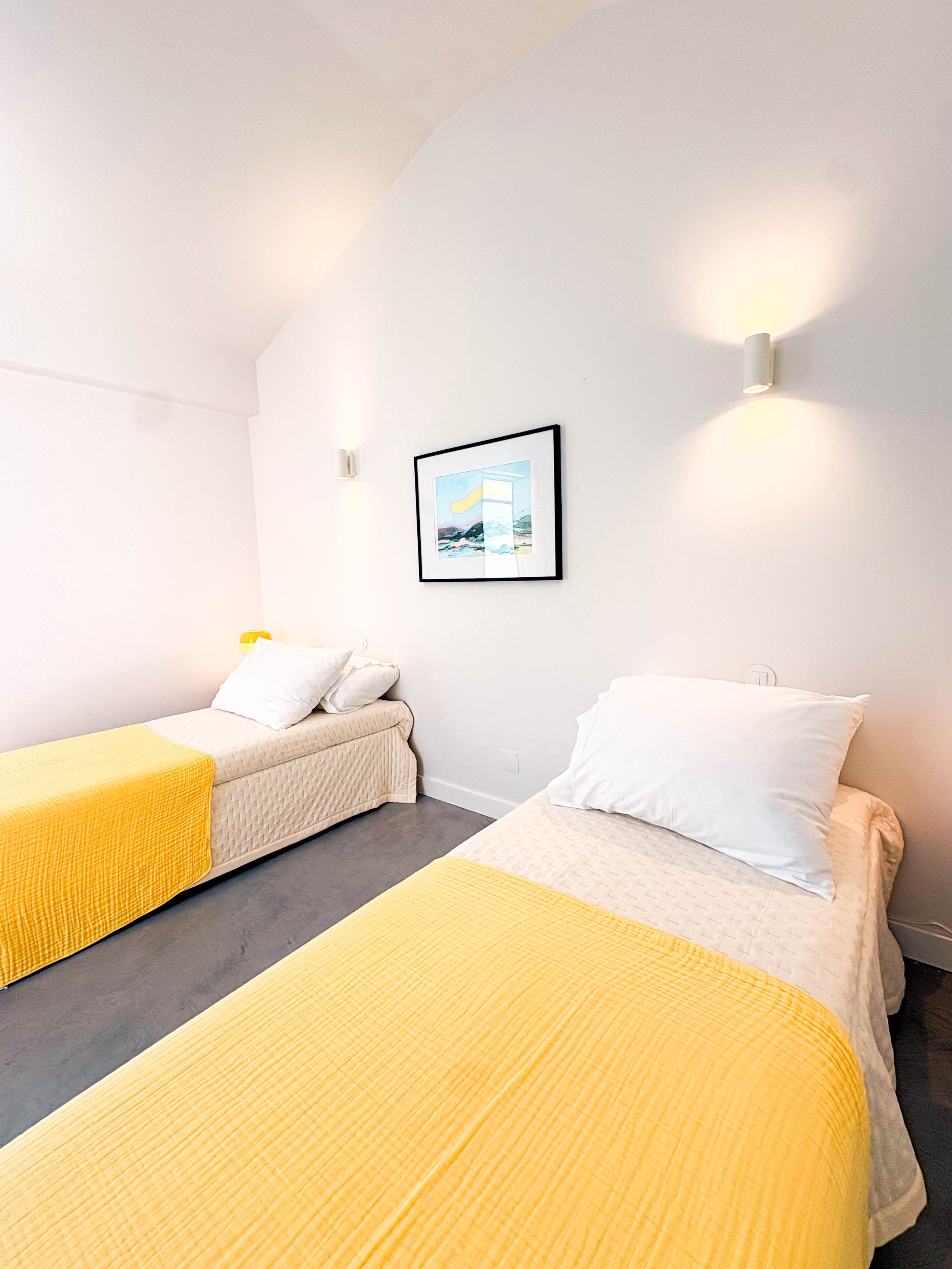 Quarto com duas camas de solteiro com roupa de cama branca e mantas amarelas, uma parede branca com uma moldura com uma foto, e duas luminárias modernas na parede