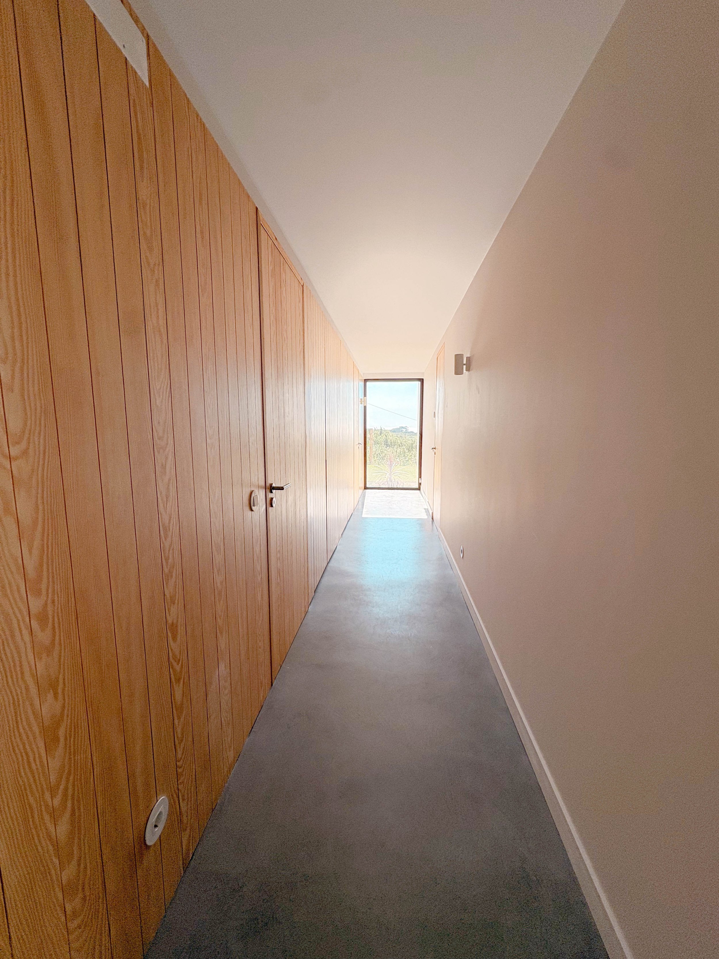 Corredor com paredes de madeira à esquerda, parede branca à direita, janela na extremidade, piso de concreto, iluminação natural.