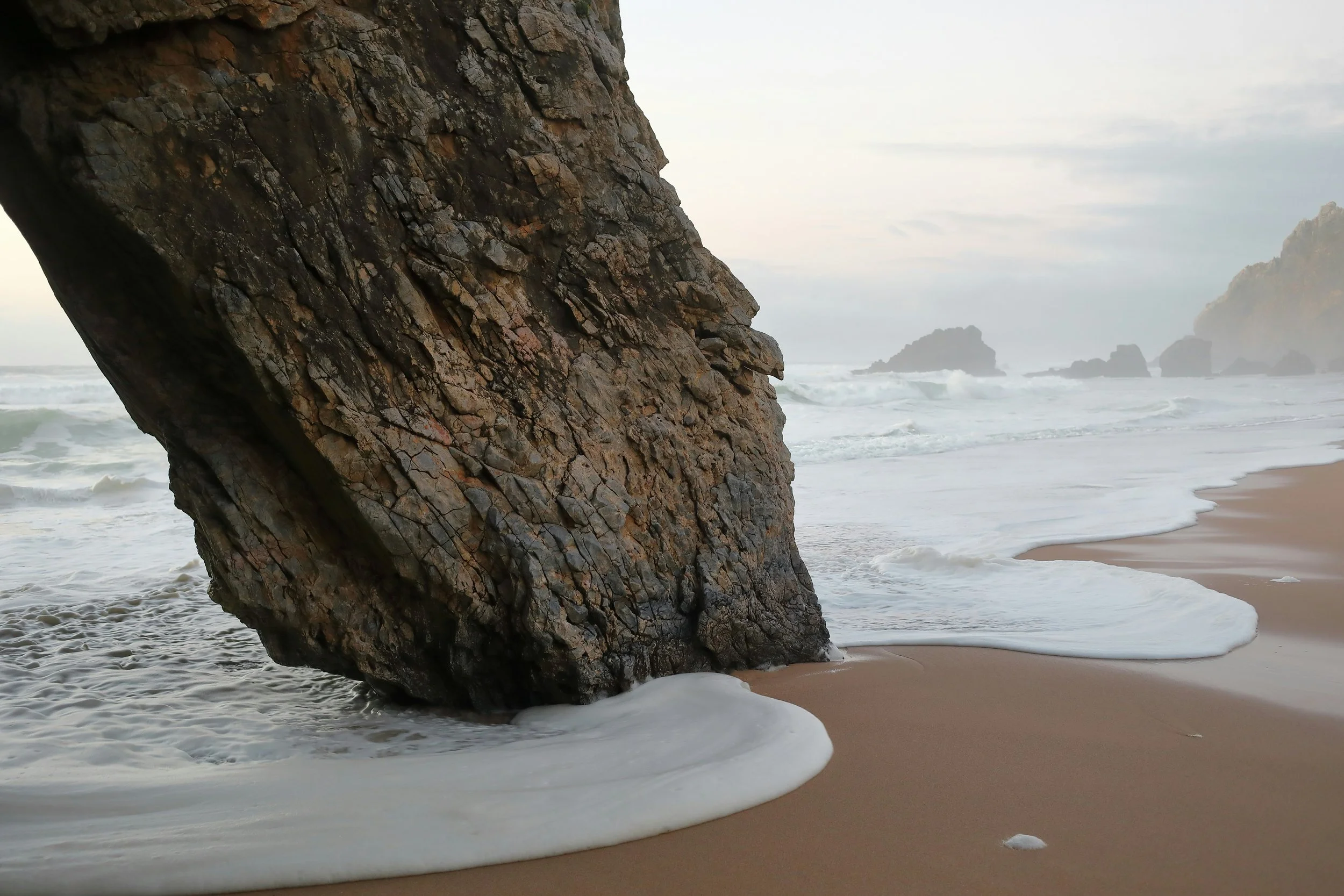 Pedra grande na praia com ondas quebrando na areia e rochas distantes no horizonte