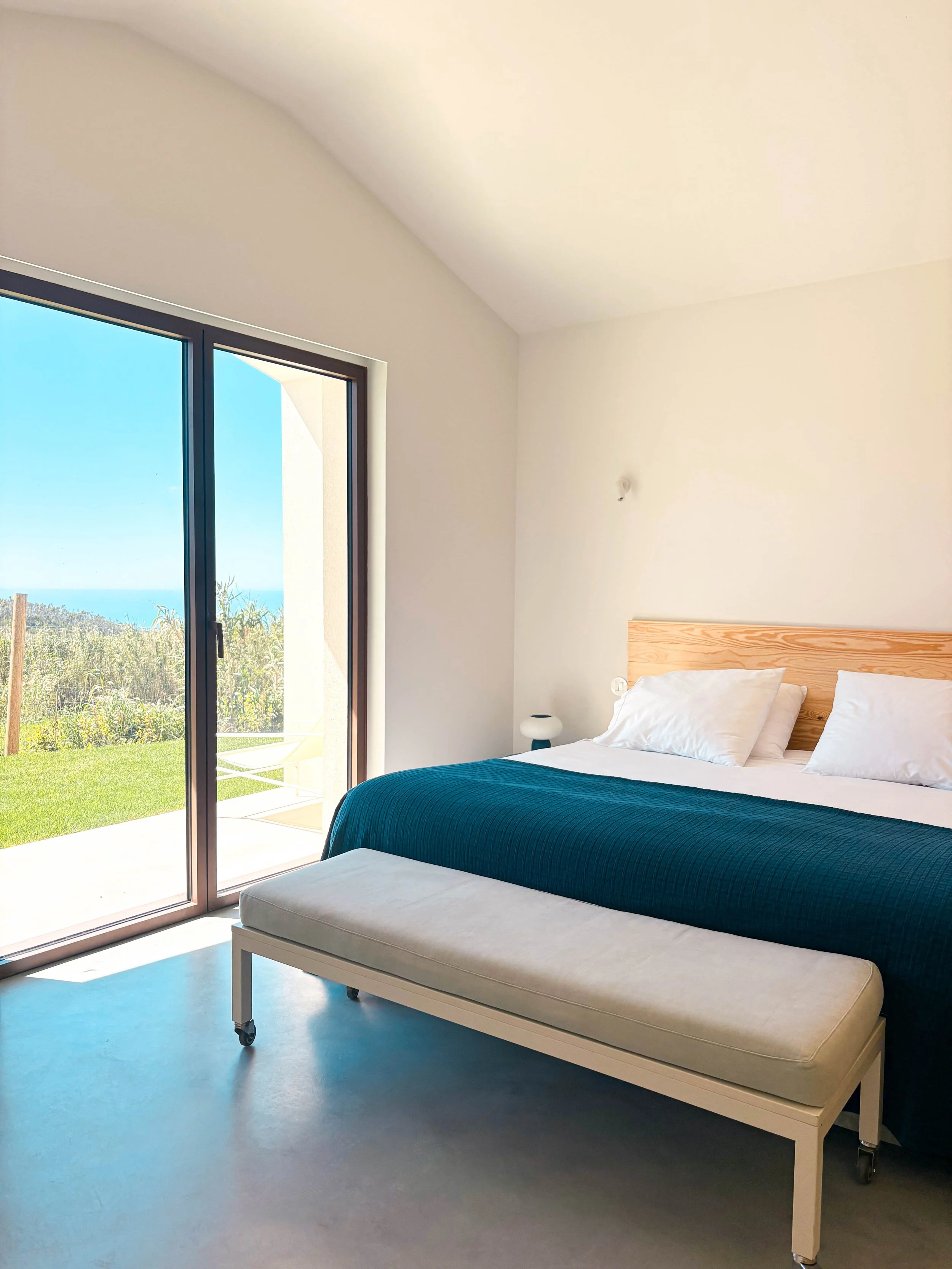 Quarto com cama de casal com cabeceira de madeira, roupa de cama branca, cobertor azul escuro, sofá pequeno ao pé da cama, e varanda com vista para o jardim e o mar ao fundo.