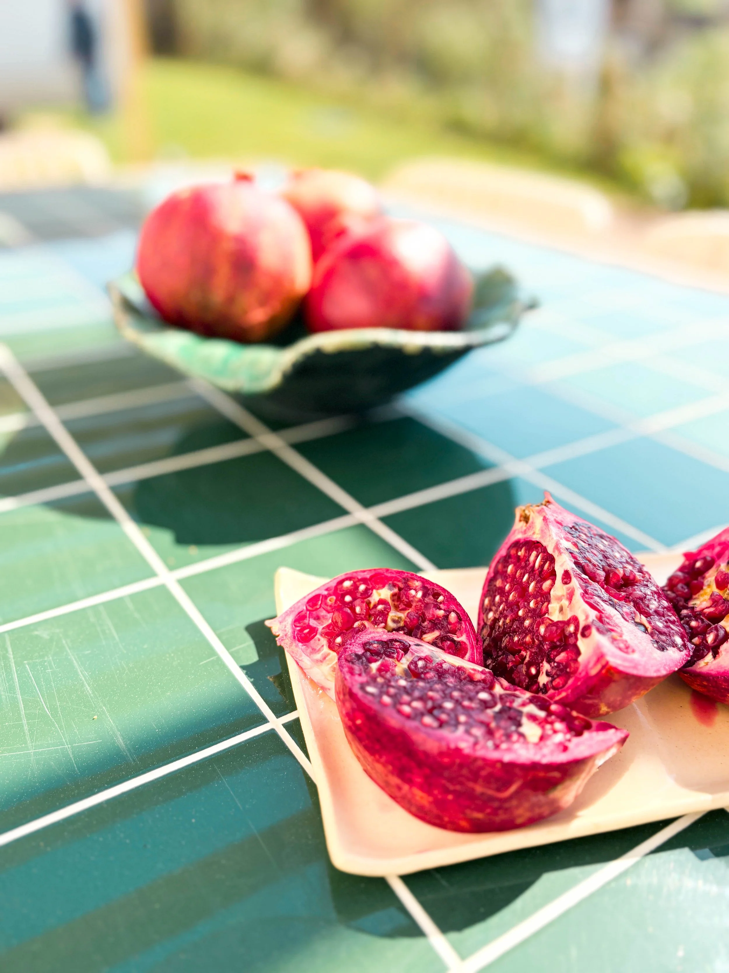 Uvas vermelhas frescas em uma tigela ao fundo e um romã cortada ao meio na frente sobre uma mesa de azulejos verdes ao ar livre.