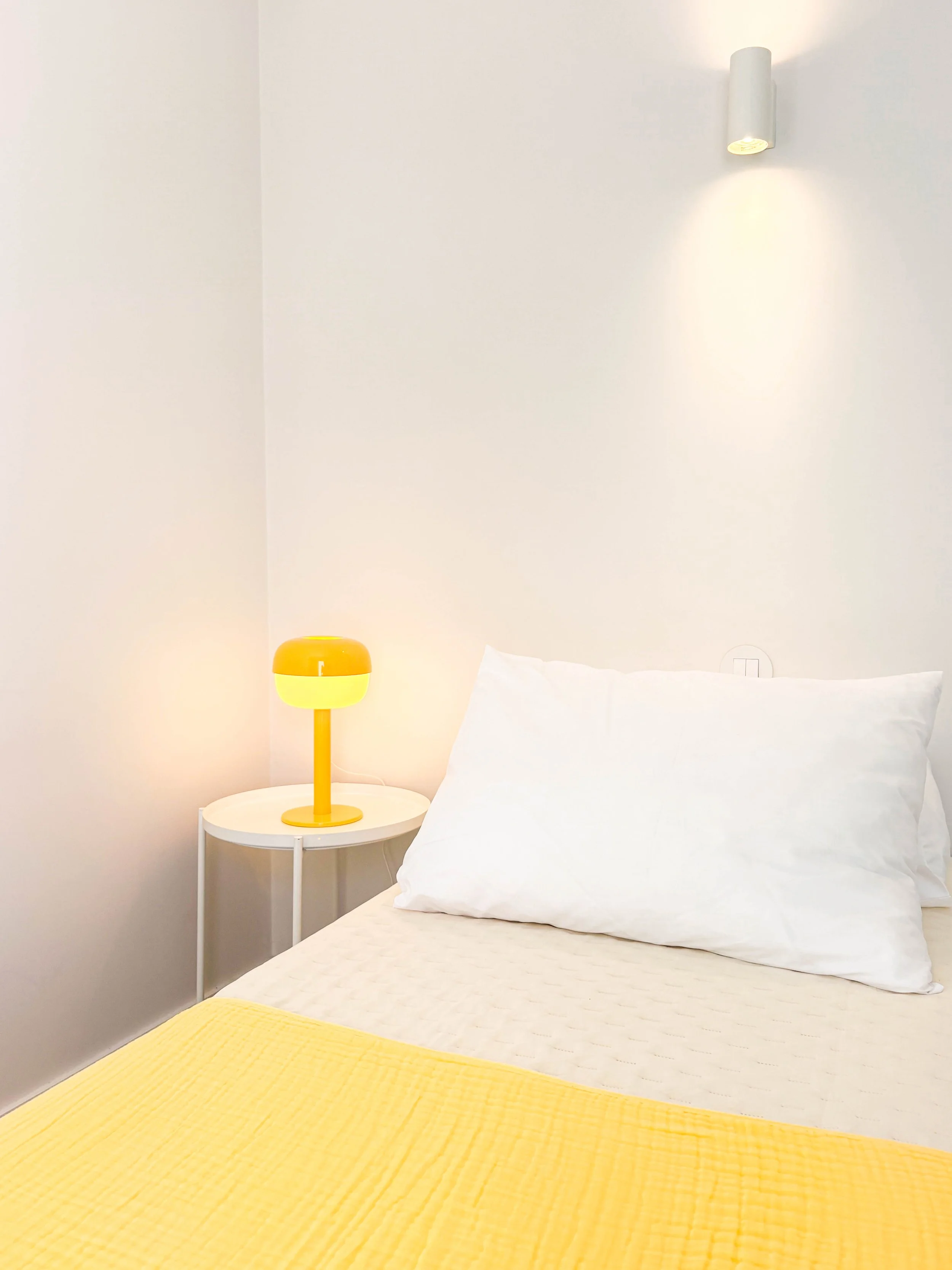 Quarto com parede branca, cama com roupa de cama branca e travesseiros brancos, edredom amarelo, luminária amarela no criado mudo ao lado da cama, e luminária branca na parede acima da cabeceira.