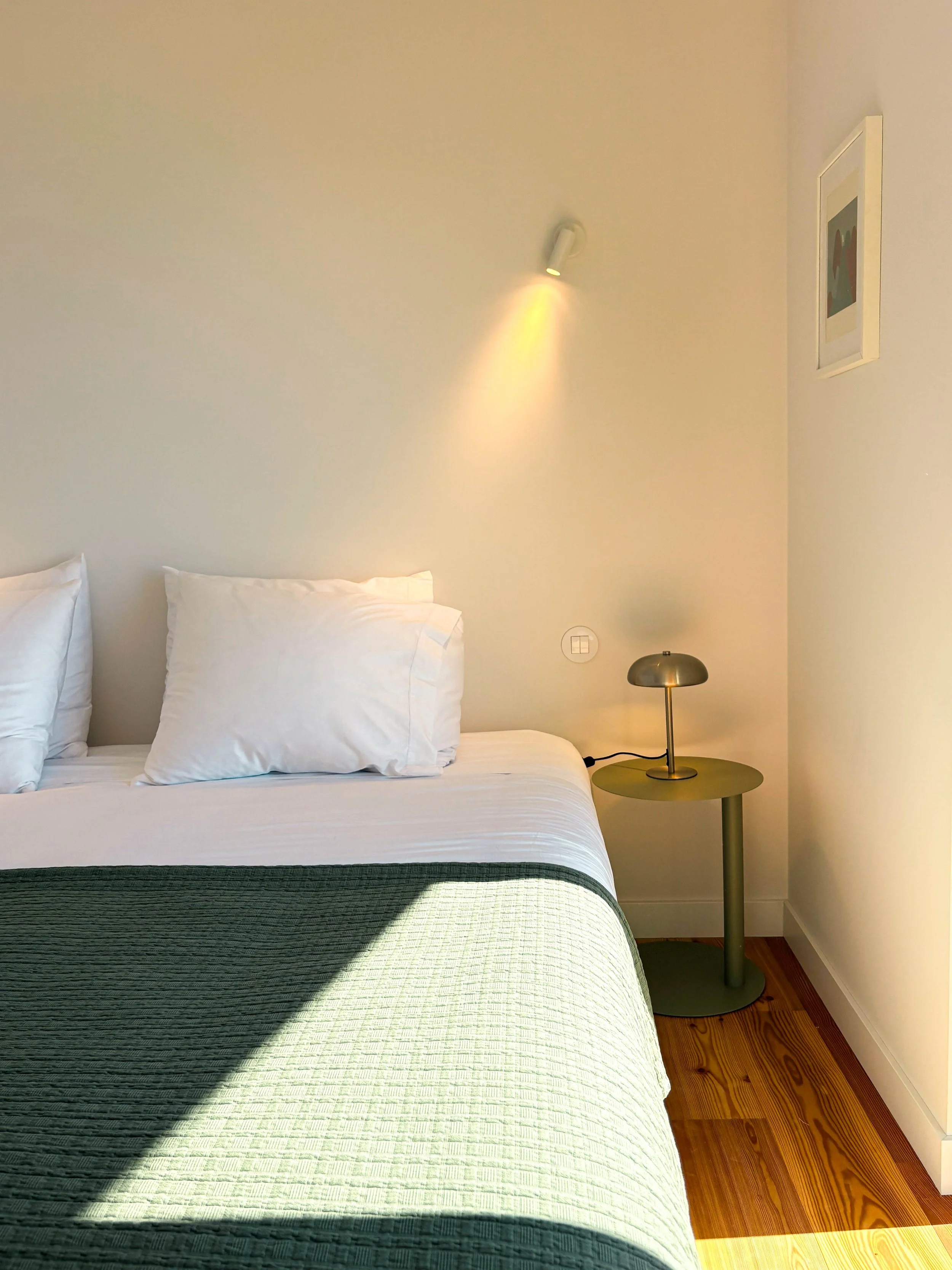 Quarto com cama de roupas brancas, travesseiros e uma colcha verde, luminária ao lado da cama, parede branca com uma foto em moldura e luz direcionada, piso de madeira.