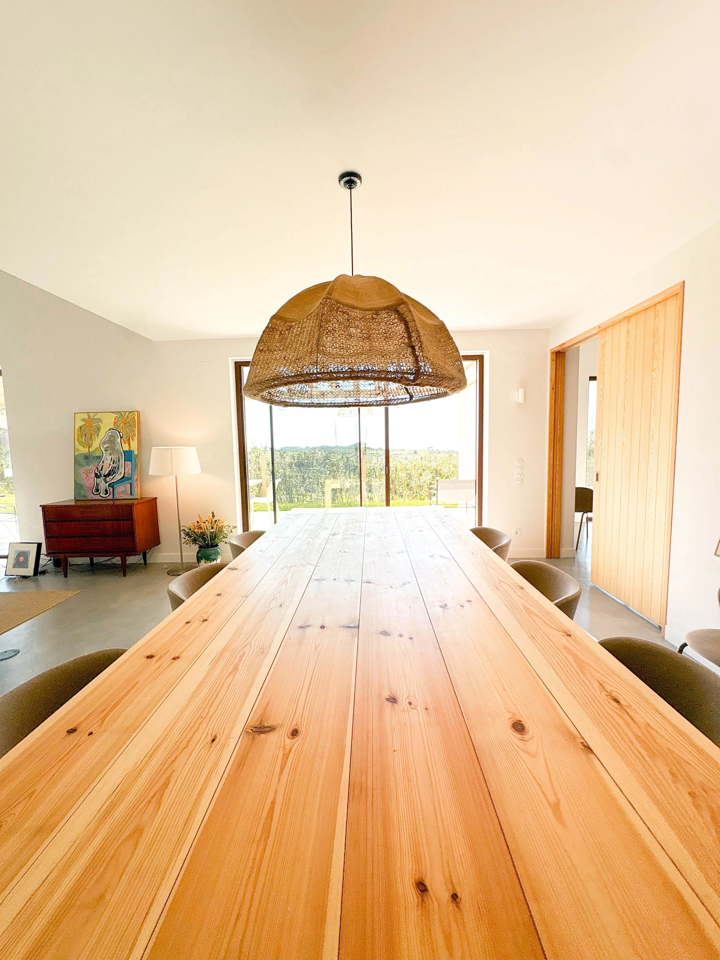 Sala de jantar com mesa de madeira longa, luz pendente de palha, janela com vista para o exterior, armário vermelho com pintura ao fundo, planta e abajur lado esquerdo.