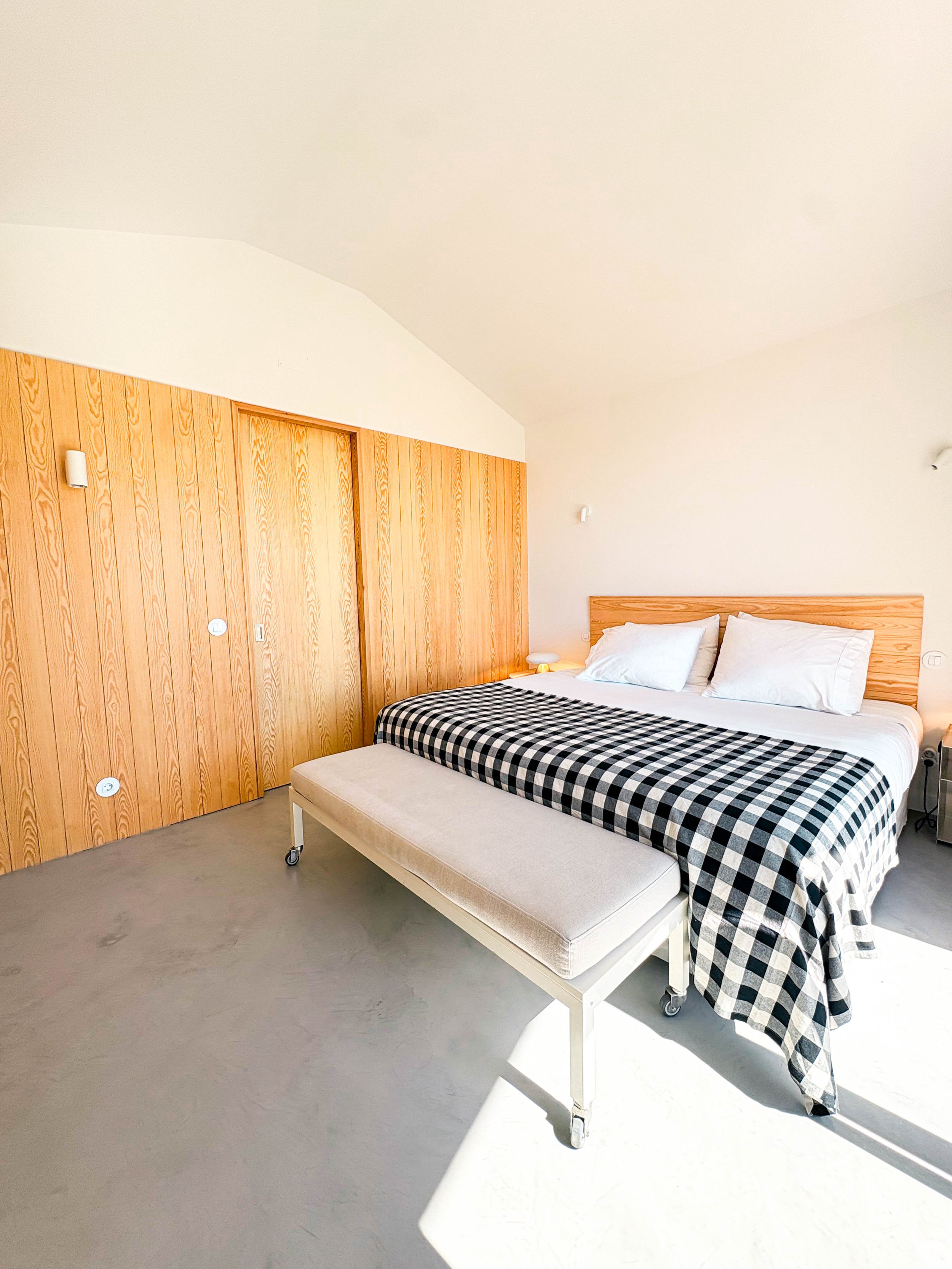 Quarto com cama de casal, almofadas brancas, cabeceira de madeira, painel de madeira na parede, e um banco branco ao pé da cama. Piso de cimento, parede branca e iluminação natural.