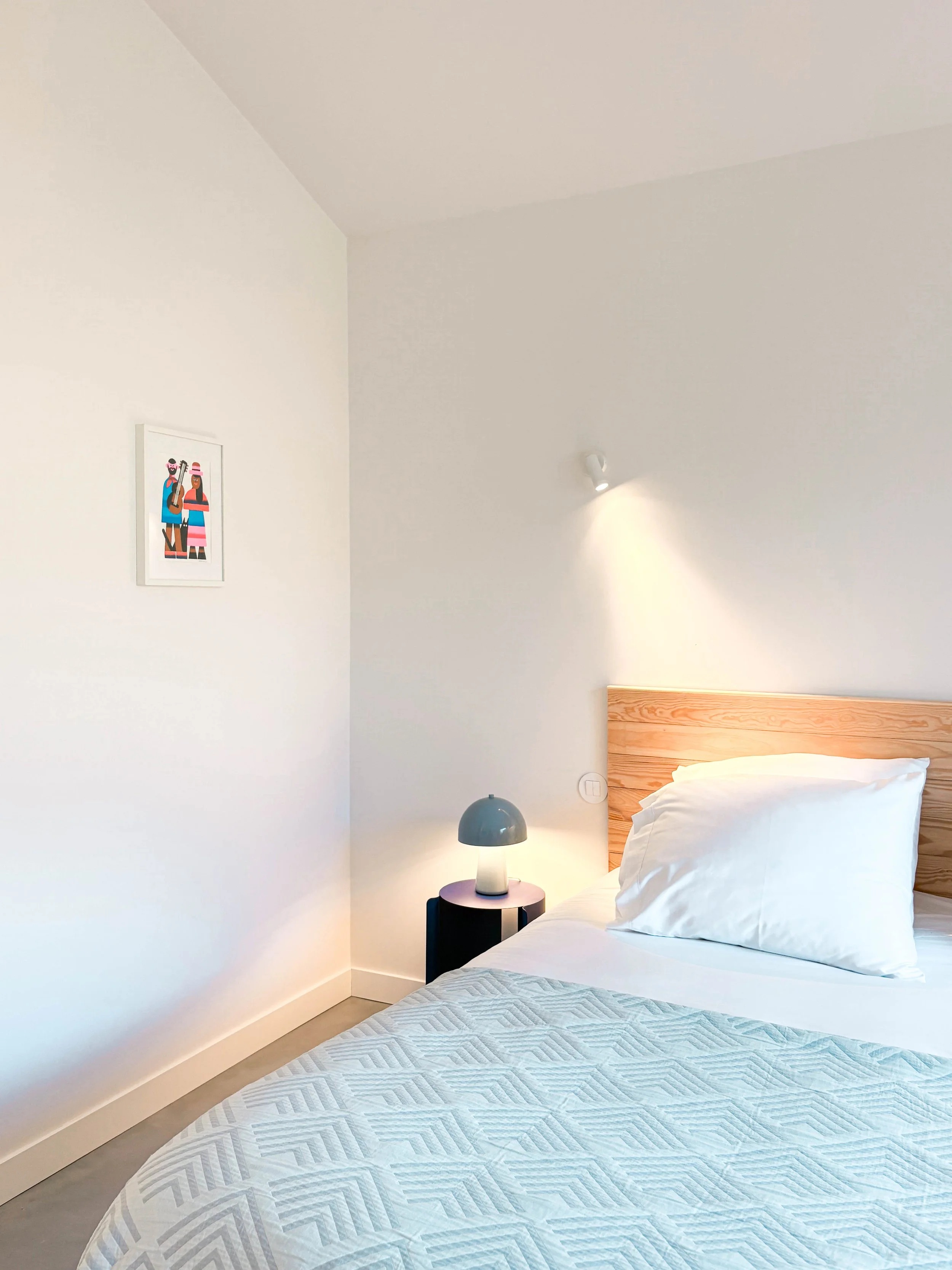 Quarto com cama de cabeceira de madeira clara, roupa de cama branca, abajur azul em mesas de canto, parede branca com uma luz direcionada e uma pintura de duas pessoas na parede ao lado.