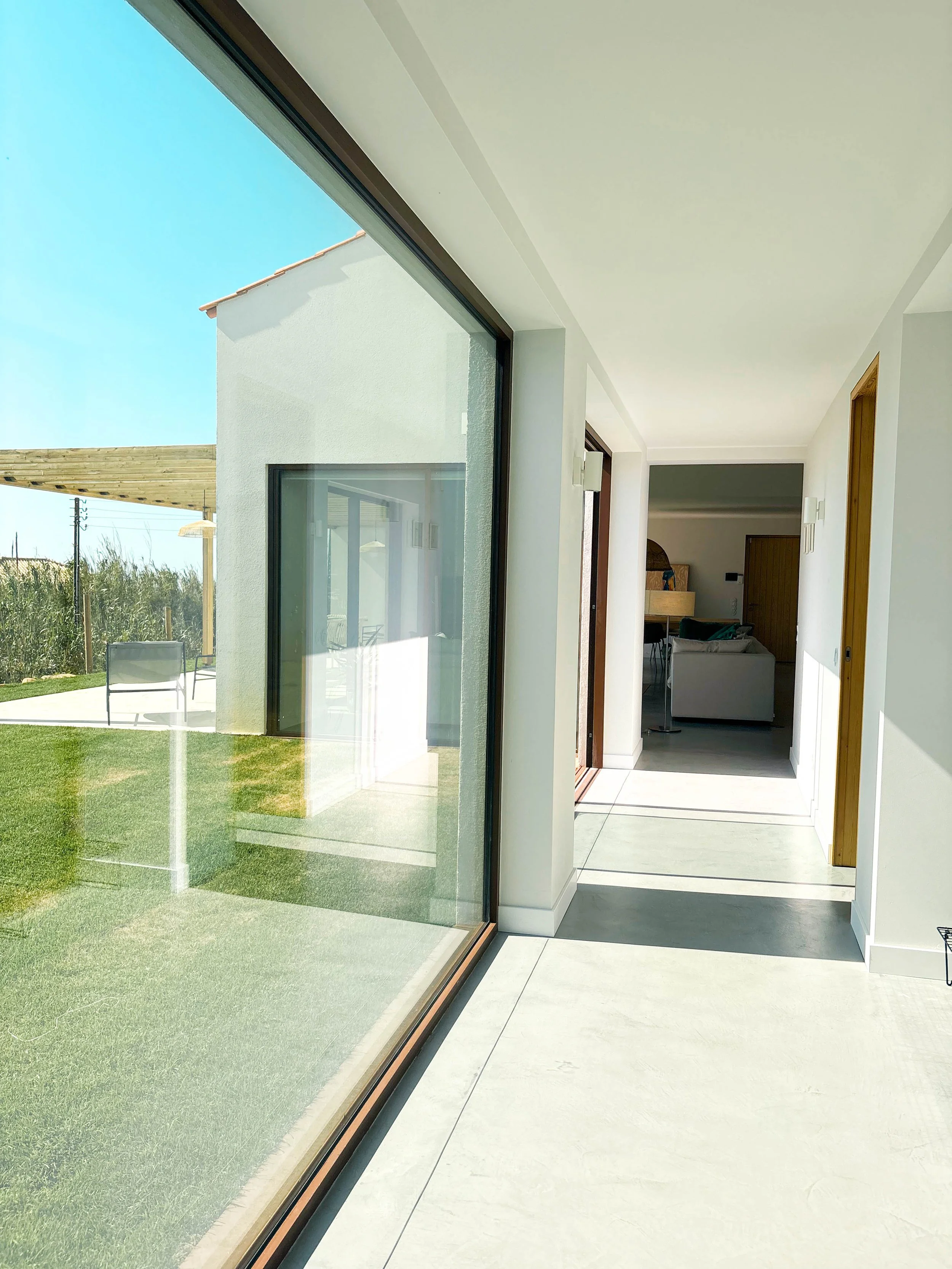 Corredor interno de uma casa com piso de concreto, paredes brancas, portas de madeira, uma janela de vidro grande que permite a entrada de luz natural, mirando um jardim externo com grama e móveis ao ar livre.