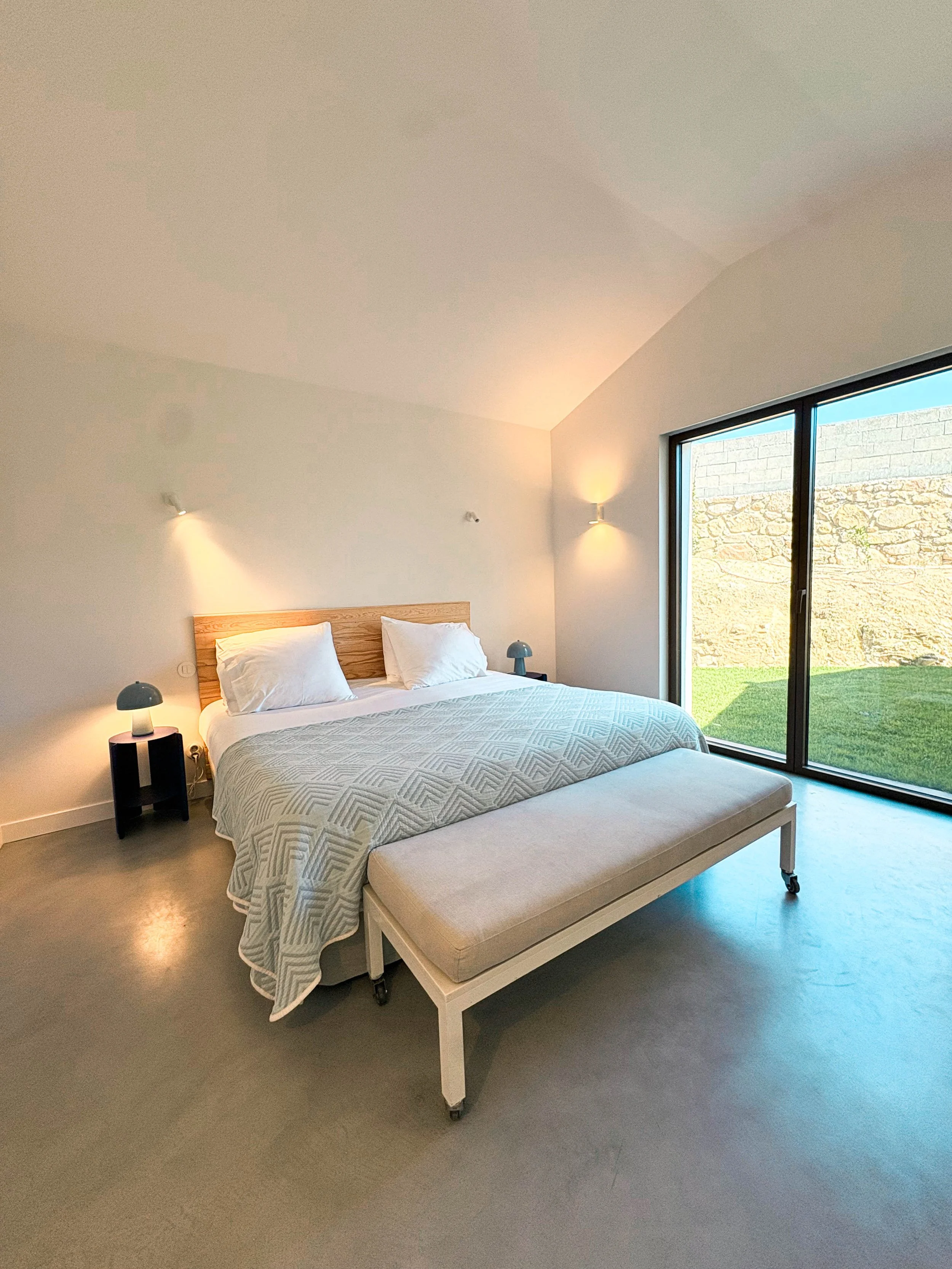 Quarto com cama de casal, dois criados-mudos com luminárias, uma langa ao lado esquerdo e outra ao lado direito da cama, uma confortová na ponta da cama, uma janela grande de vidro com vista para um jardim gramado e uma parede de pedra ao fundo.