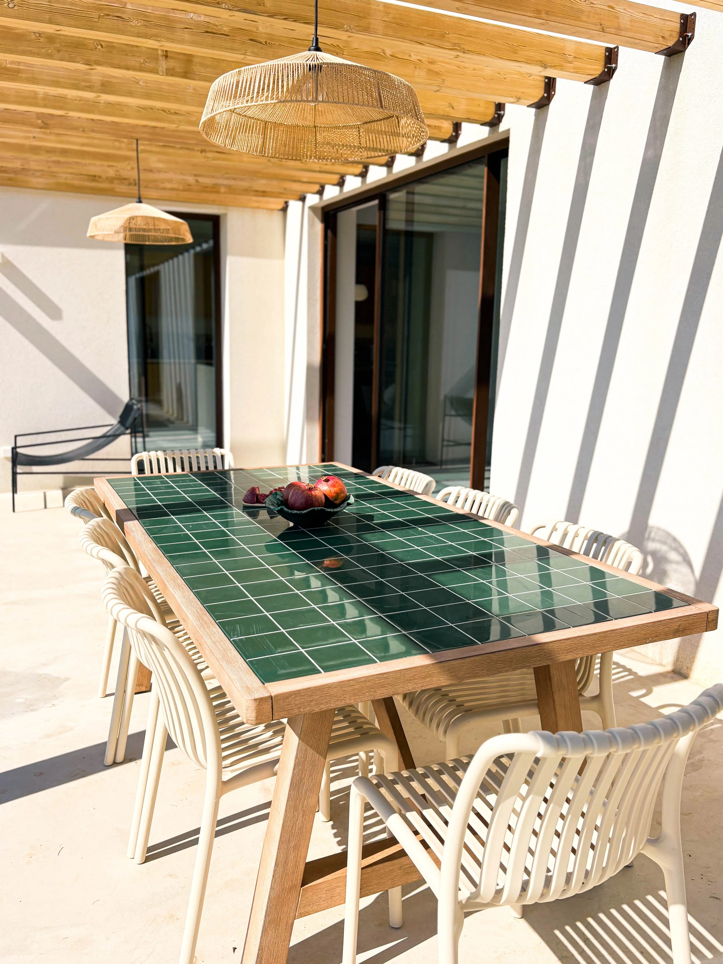 Mesa de jantar com tampo de azulejos verdes, cercada por cadeiras brancas, em um espaço ao ar livre. Acima, pendem duas luminárias de fibra natural. No fundo, há uma parede branca, uma porta de vidro e uma janela, com a sombra das luminárias projetad