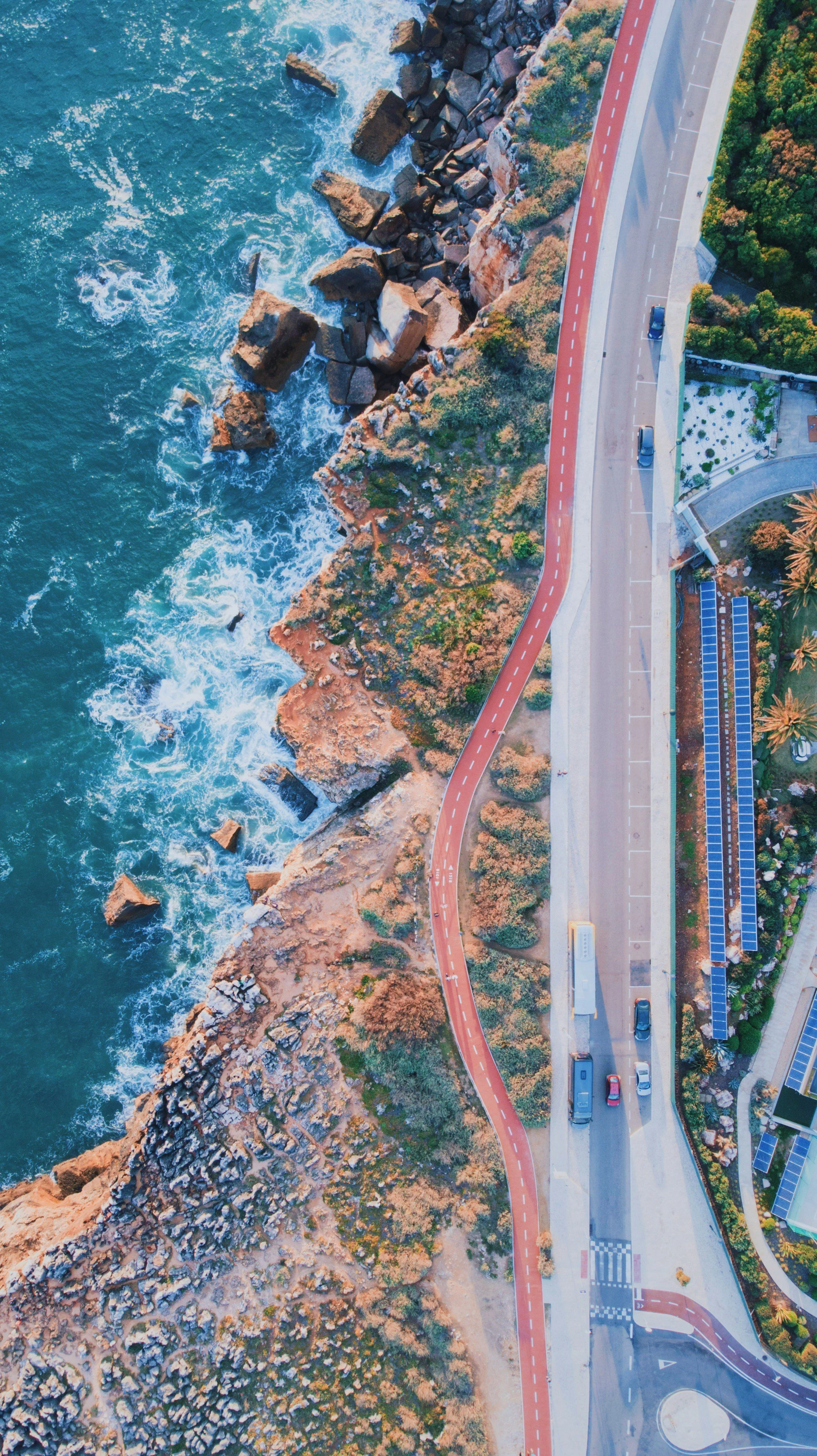 Imagem aérea de uma estrada ao lado de um litoral rochoso, com o mar ao lado esquerdo e vegetação no lado direito.