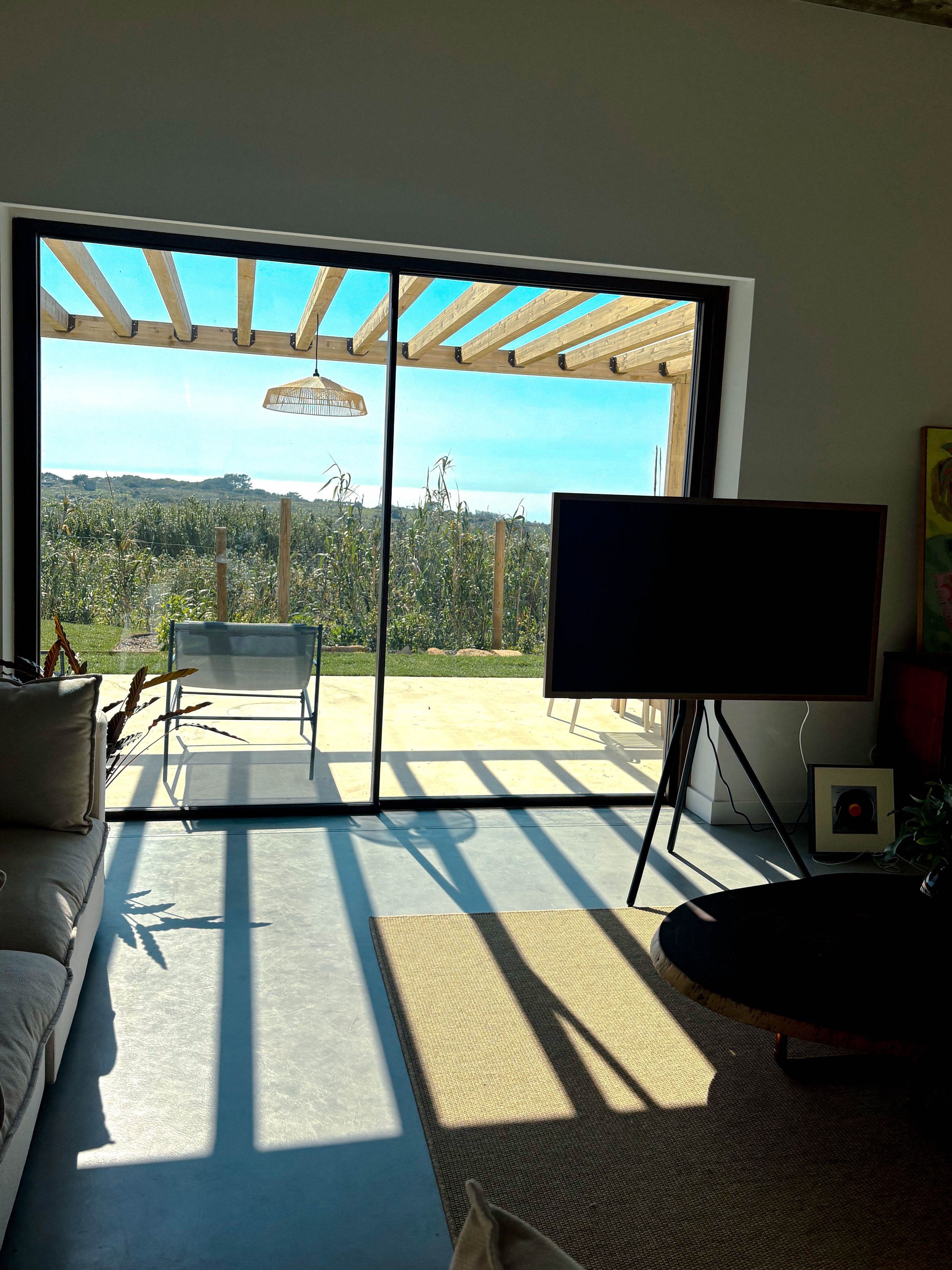 Sala de estar com um grande portal de vidro que dá para uma varanda com jardim e árvores ao fundo, deixando a luz natural entrar. Há uma televisão na sala e uma cadeira na varanda ao ar livre.