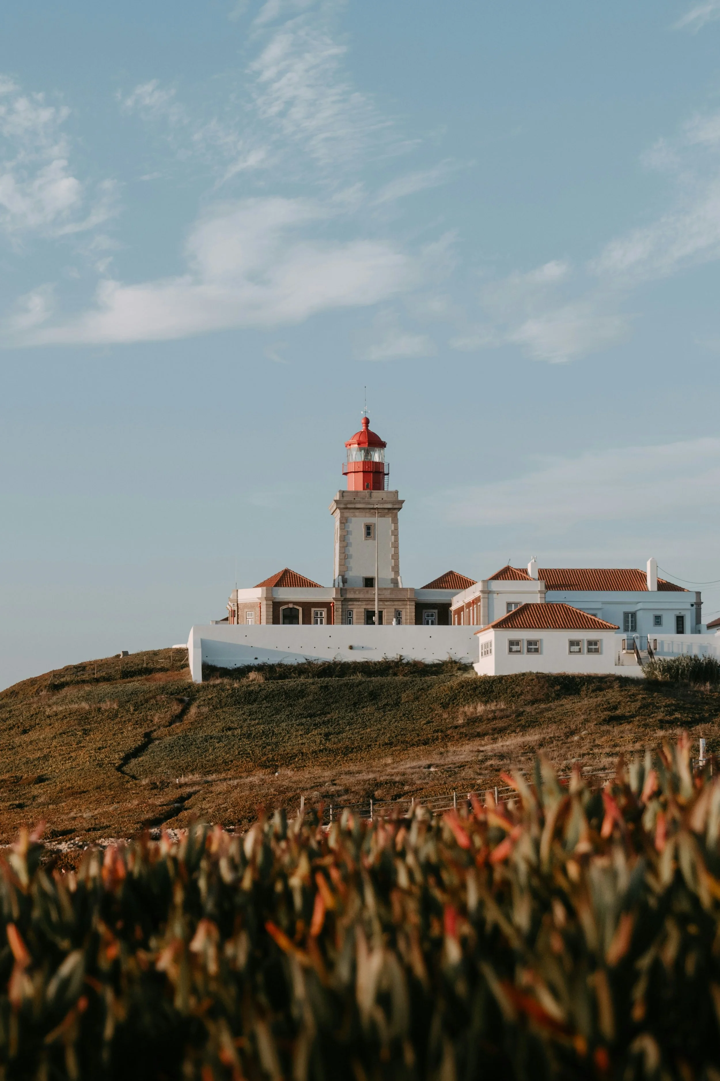 Faro com edifícios brancos e telhados vermelhos situado em uma colina, sob céu azul com nuvens leves.
