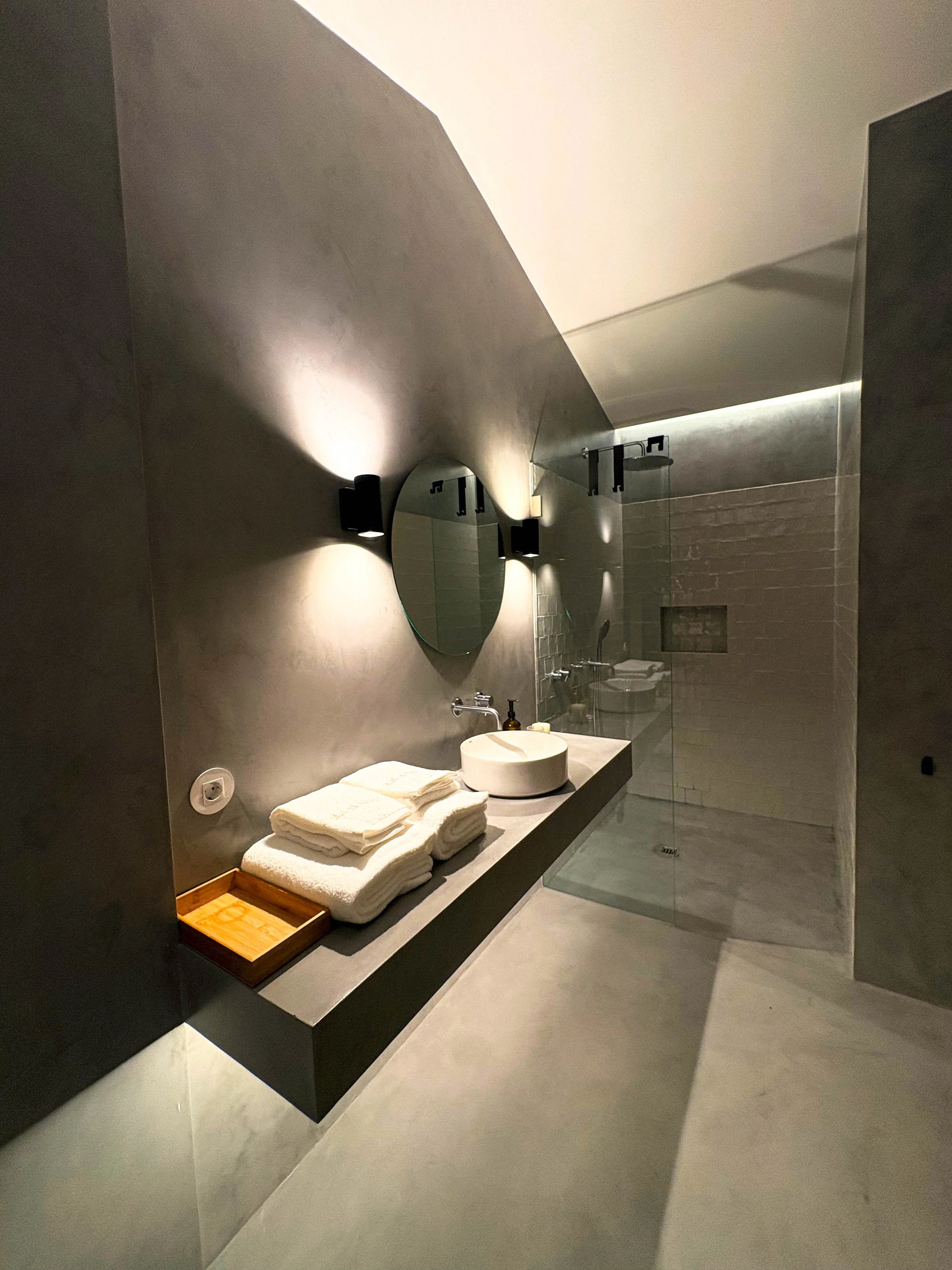 Banheiro moderno com espelho redondo, parede de cimento, iluminação de parede, pia de vaso sanitário no balcão, toalhas brancas empilhadas em uma bandeja de madeira, cabine de vidro com piso de cimento.