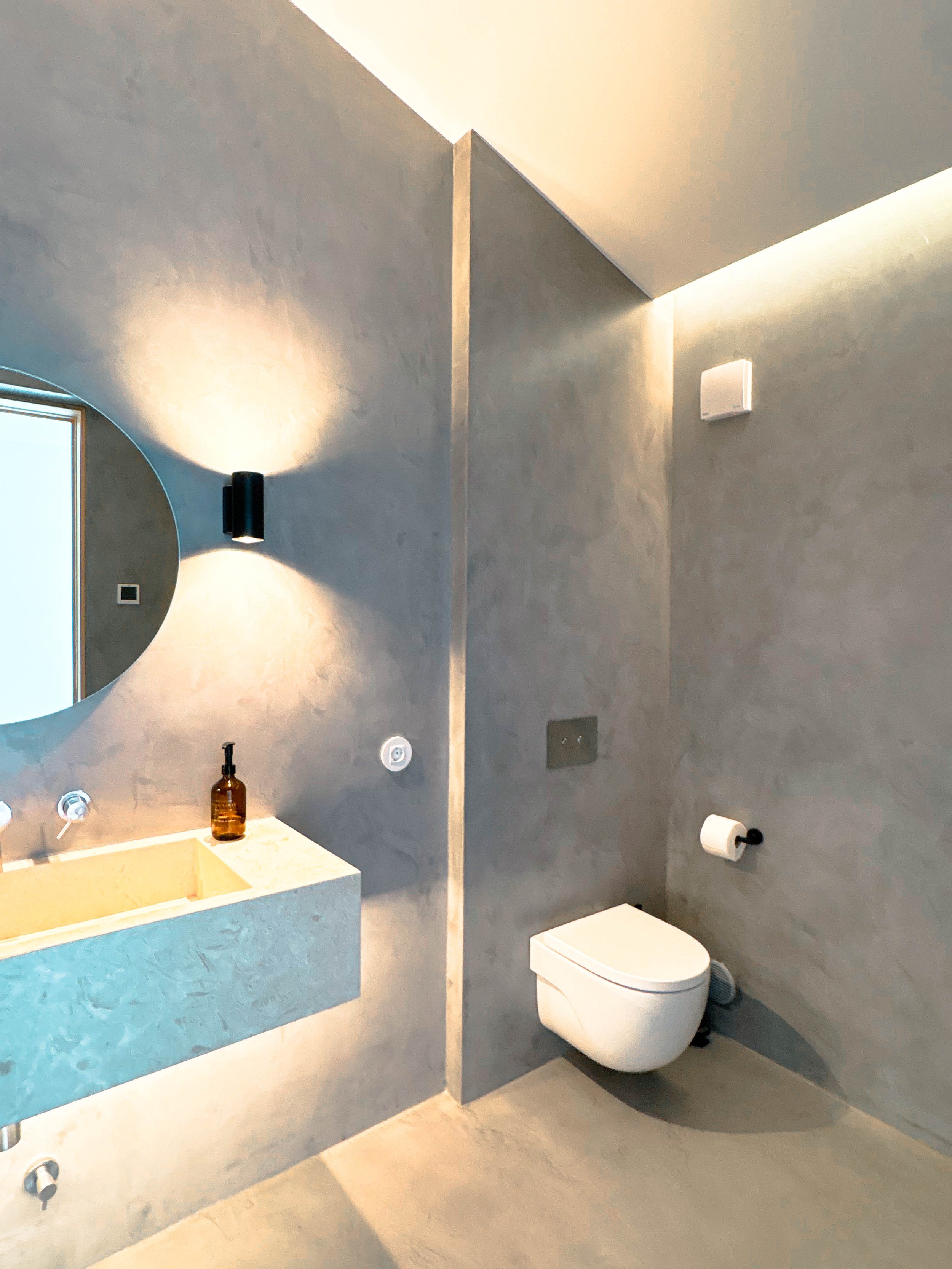 Banheiro moderno com espelho, pia de pedra, vaso sanitário inteligente, iluminação indireta e parede de tom cinza.