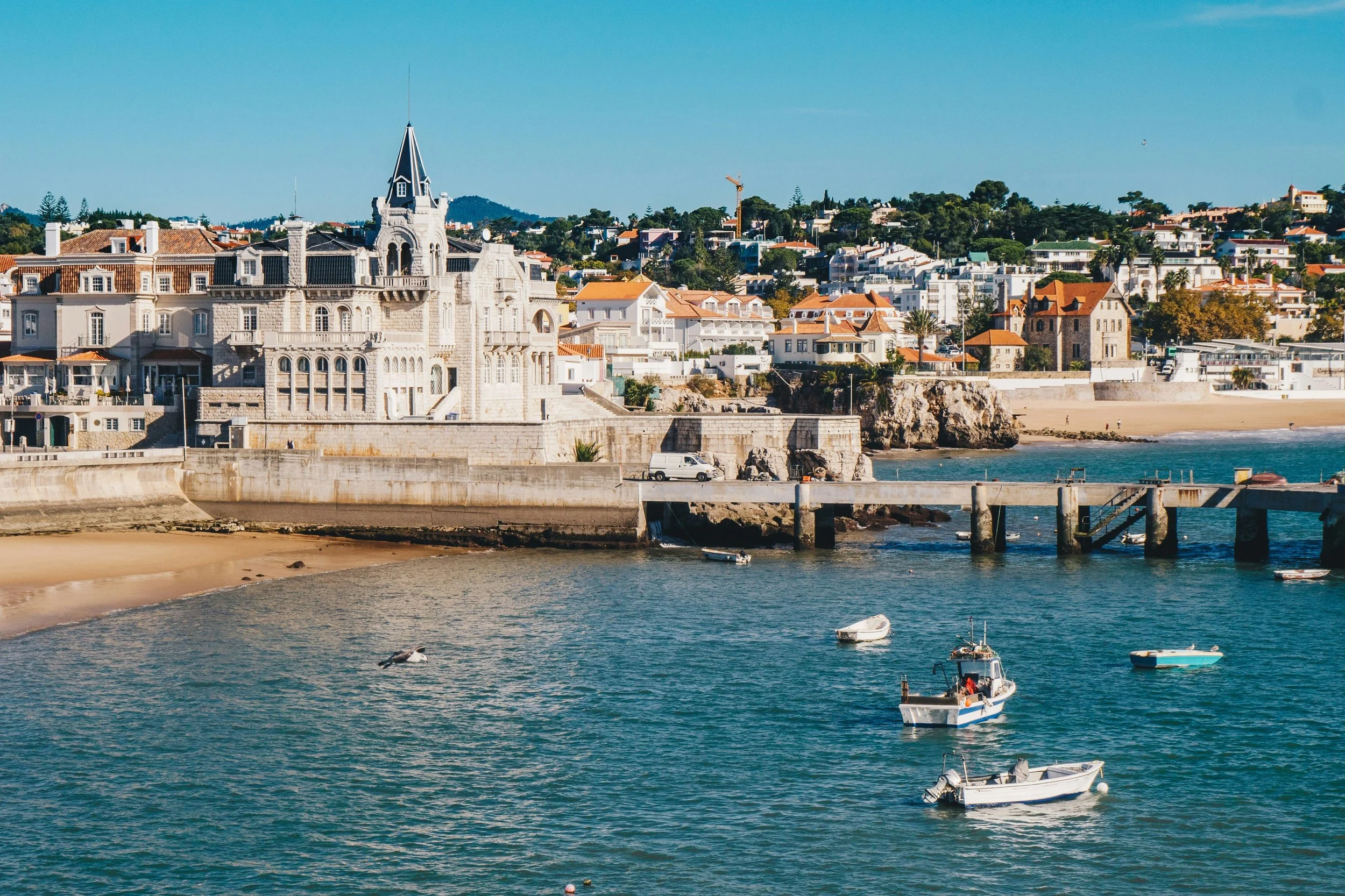 Vista de uma praia com água azul, barcos dockados próximos à praia, e uma cidade com edifícios brancos e um castelo de pedra com torre alta ao fundo.