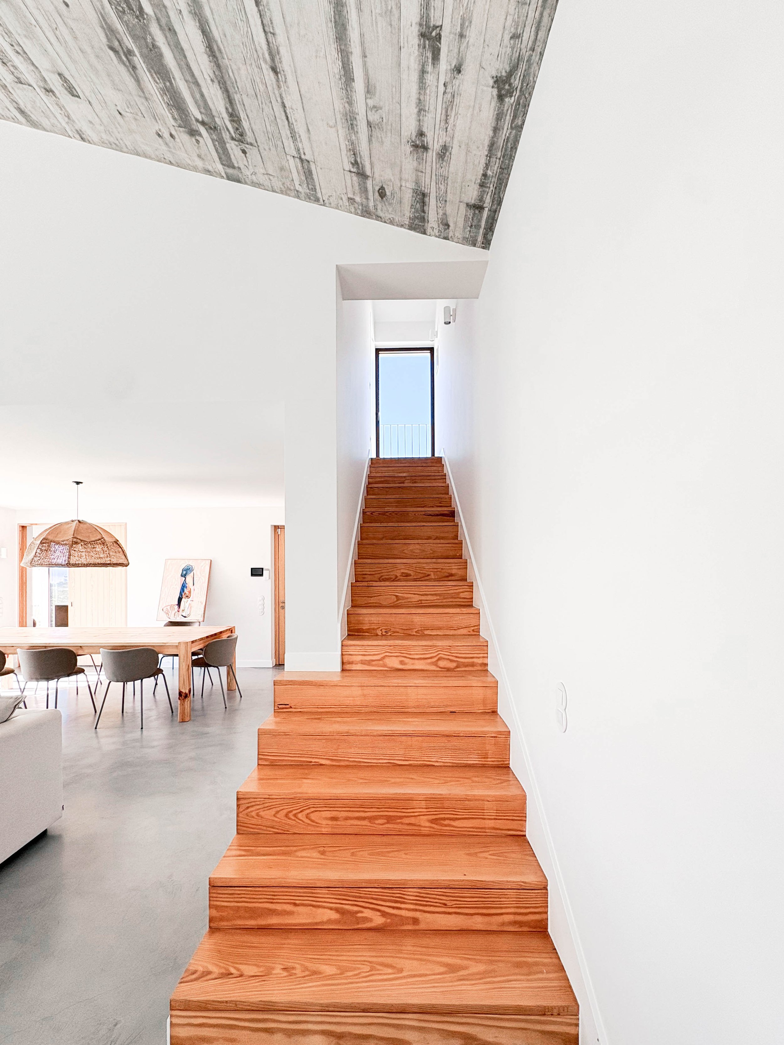 Escada de madeira com degraus, lado direito, parede branca ao fundo e teto de concreto exposto. Sala com mesa de jantar e cadeiras visíveis à esquerda.