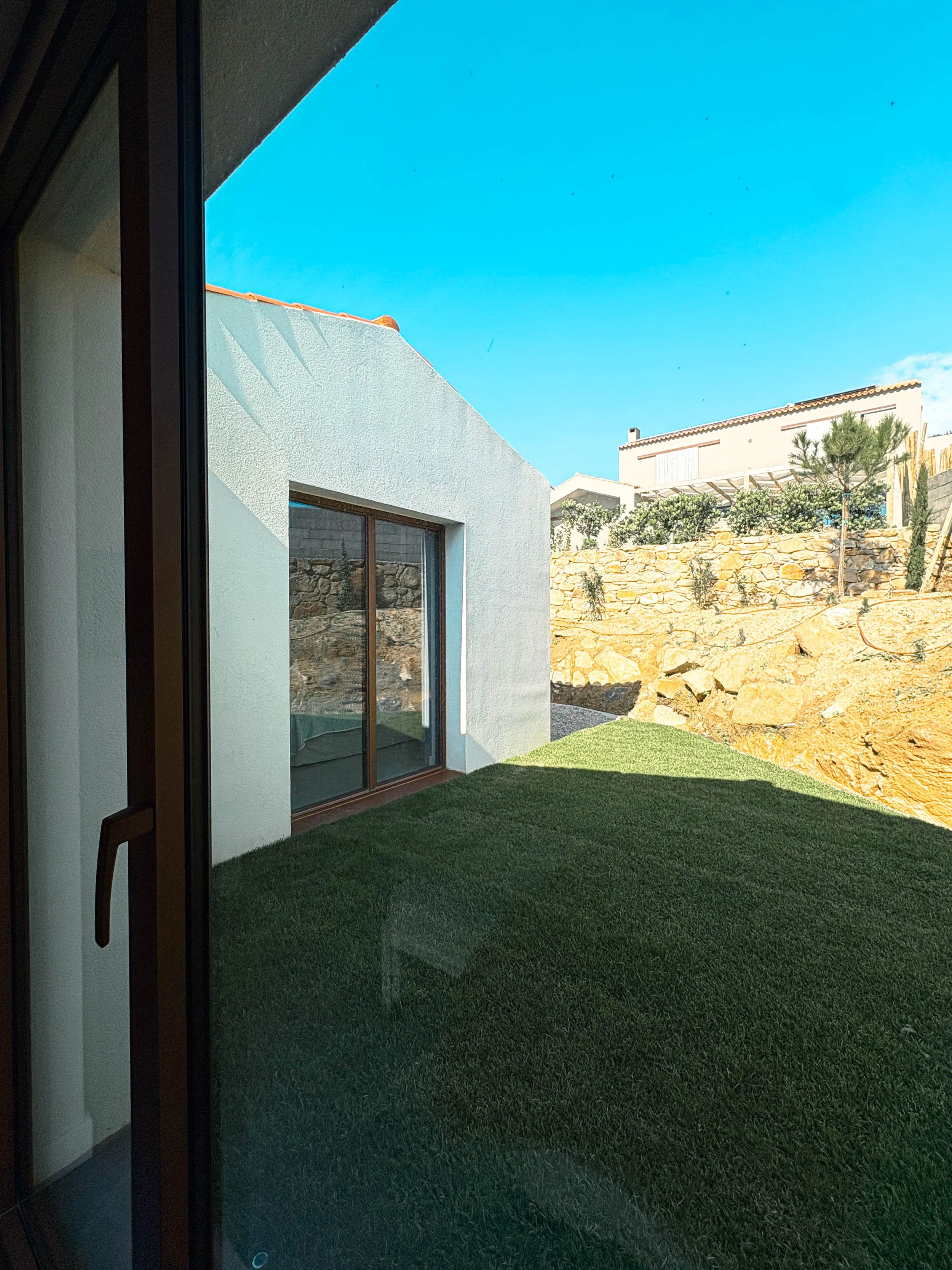 Vista de um quintal com gramado verde, paredes brancas de uma casa moderna e cerca de pedras, sob céu claro e azul.