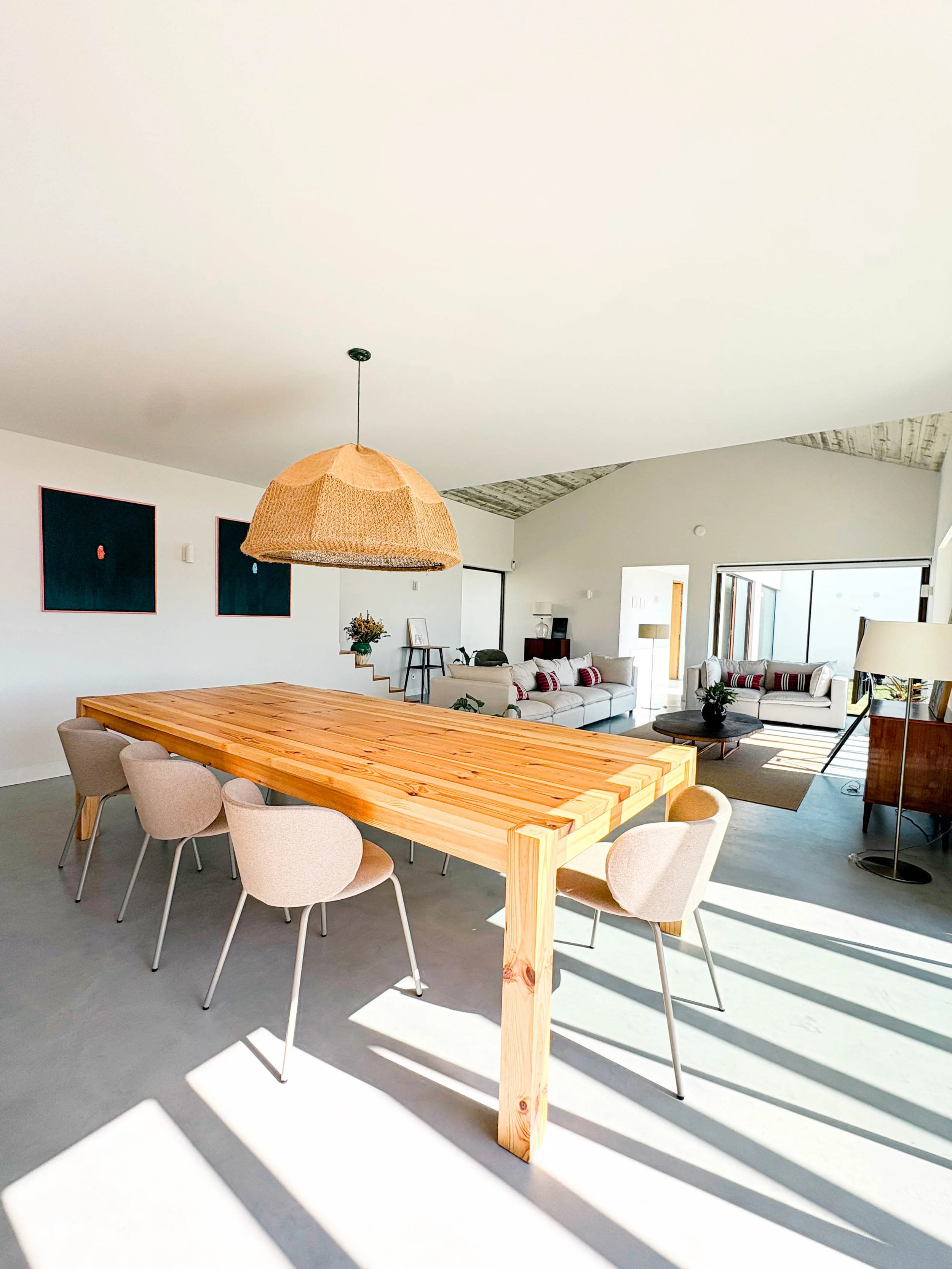 Sala de estar moderna com mesa de madeira e cadeiras beige, sofás brancos com almofadas listradas vermelhas e brancas, espaço bem iluminado com janelas grandes e decoração minimalista.