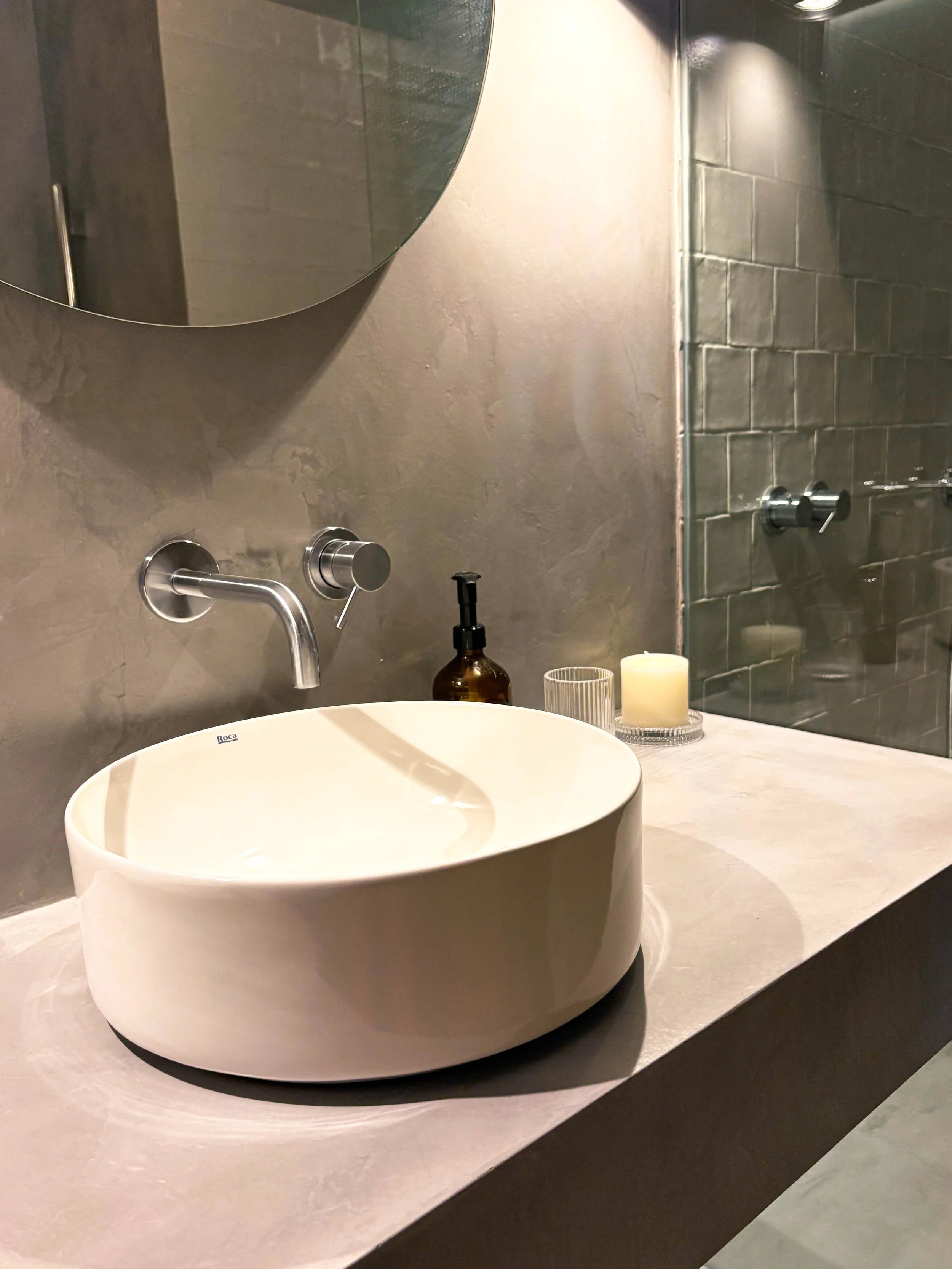 Espelho redondo, pia de vaso sanitário branco, torneira moderna, sabonete líquido em pote de vidro, vela em recipiente de vidro, parede de azulejo cinza, lavatório de concreto.