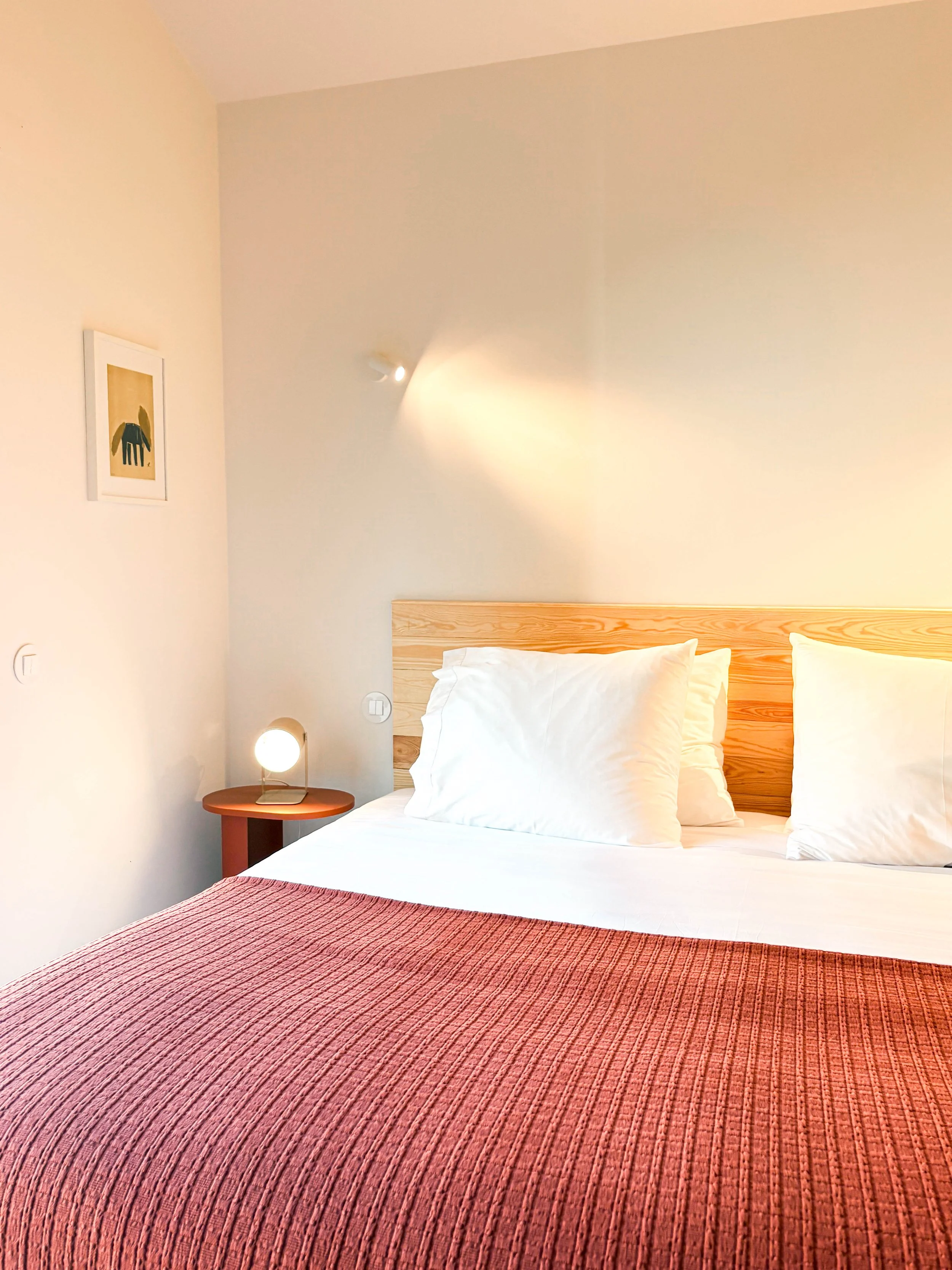 Quarto com cama de casal com cabeceira de madeira, roupas de cama brancas, uma almofada, uma luminária de mesa acesa e uma parede decorada com quadro de um elefante destacado em um ambiente de luz quente.