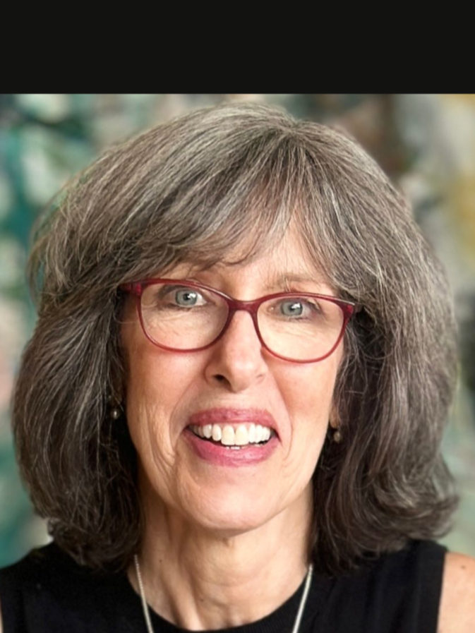 Close-up image of a smiling older woman with gray hair, glasses, and wearing a black top, standing outdoors with a blurred natural background.