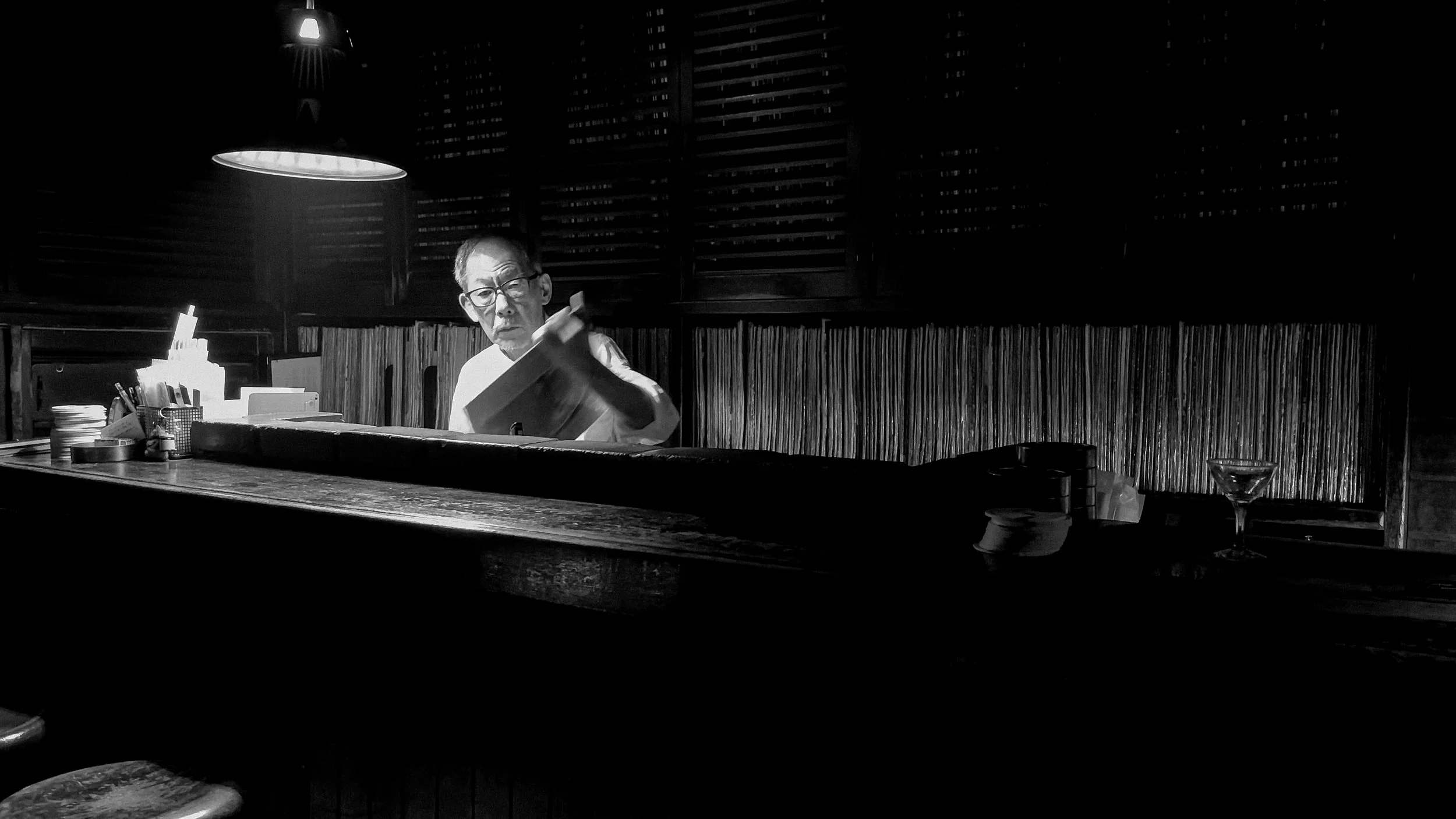 Elderly Japanese bar owner absorbed in reading beneath a single hanging lamp, Tokyo, 2024. Black and white street photography by Aidan Martin