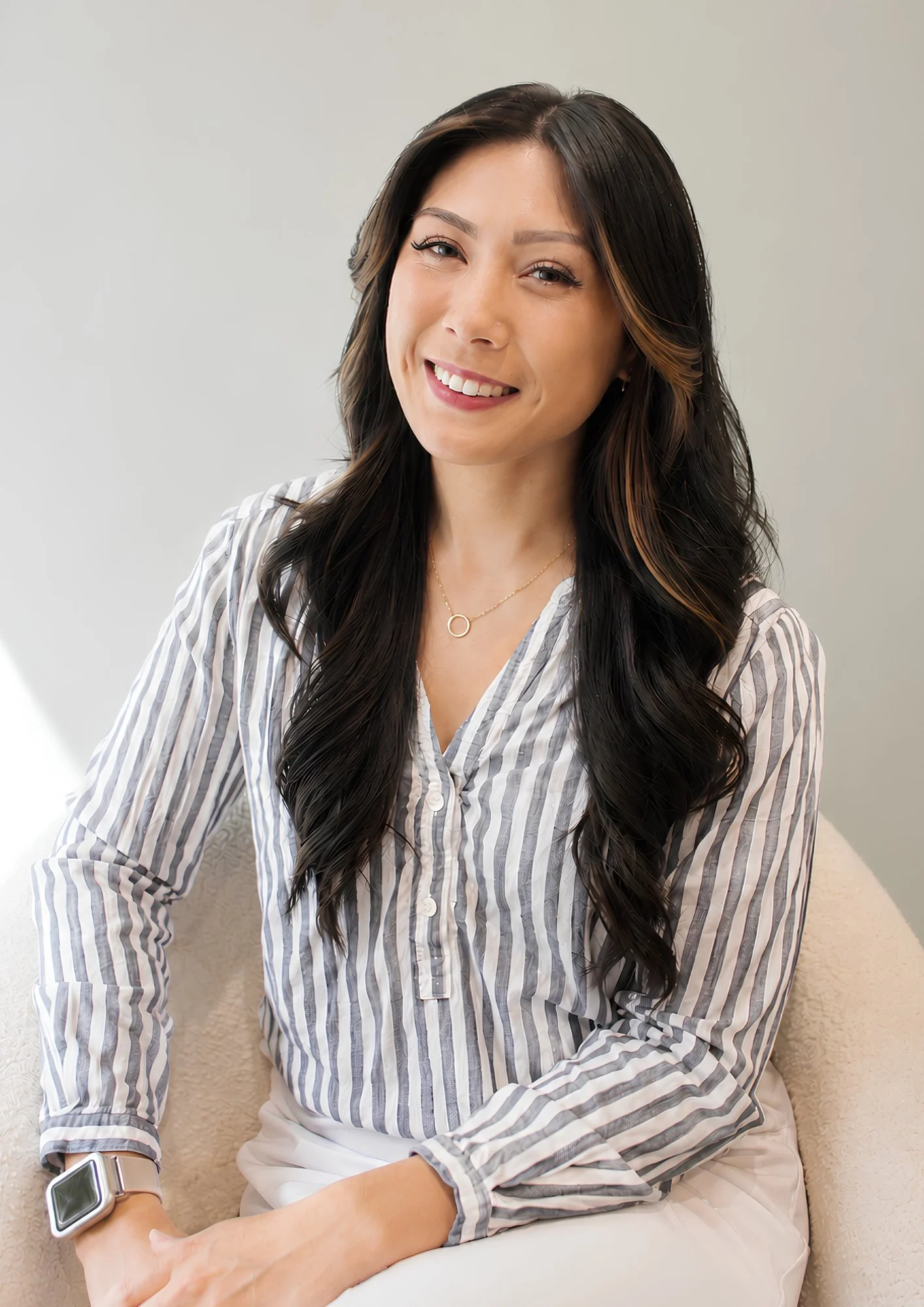 A woman with long dark wavy hair, wearing a striped shirt, sitting on a beige chair against a neutral background, smiling at the camera.