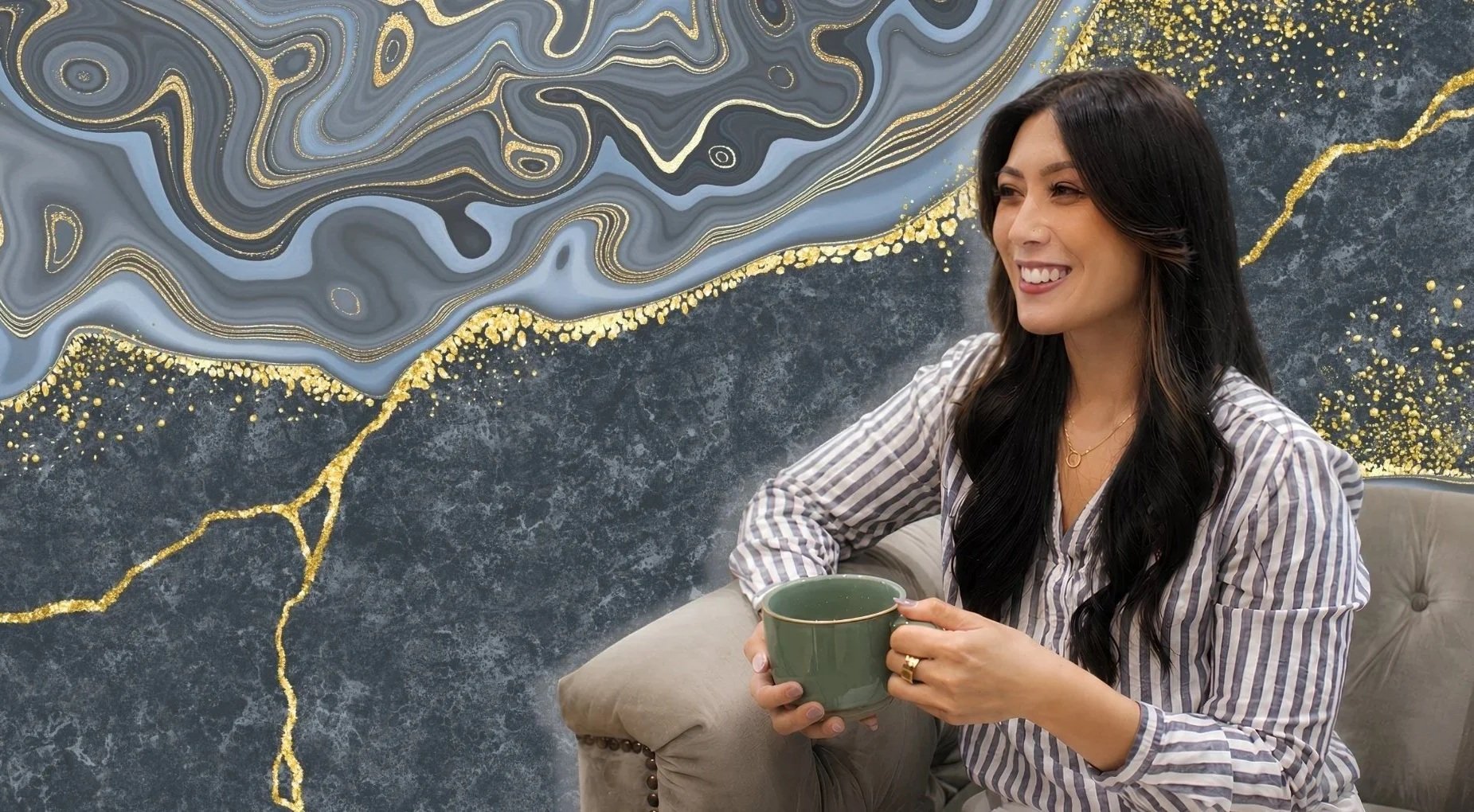 A woman with long black hair, wearing a striped shirt and a gold necklace, sitting on a beige couch and holding a green mug, smiling in front of a dark abstract marble and gold accent wall.