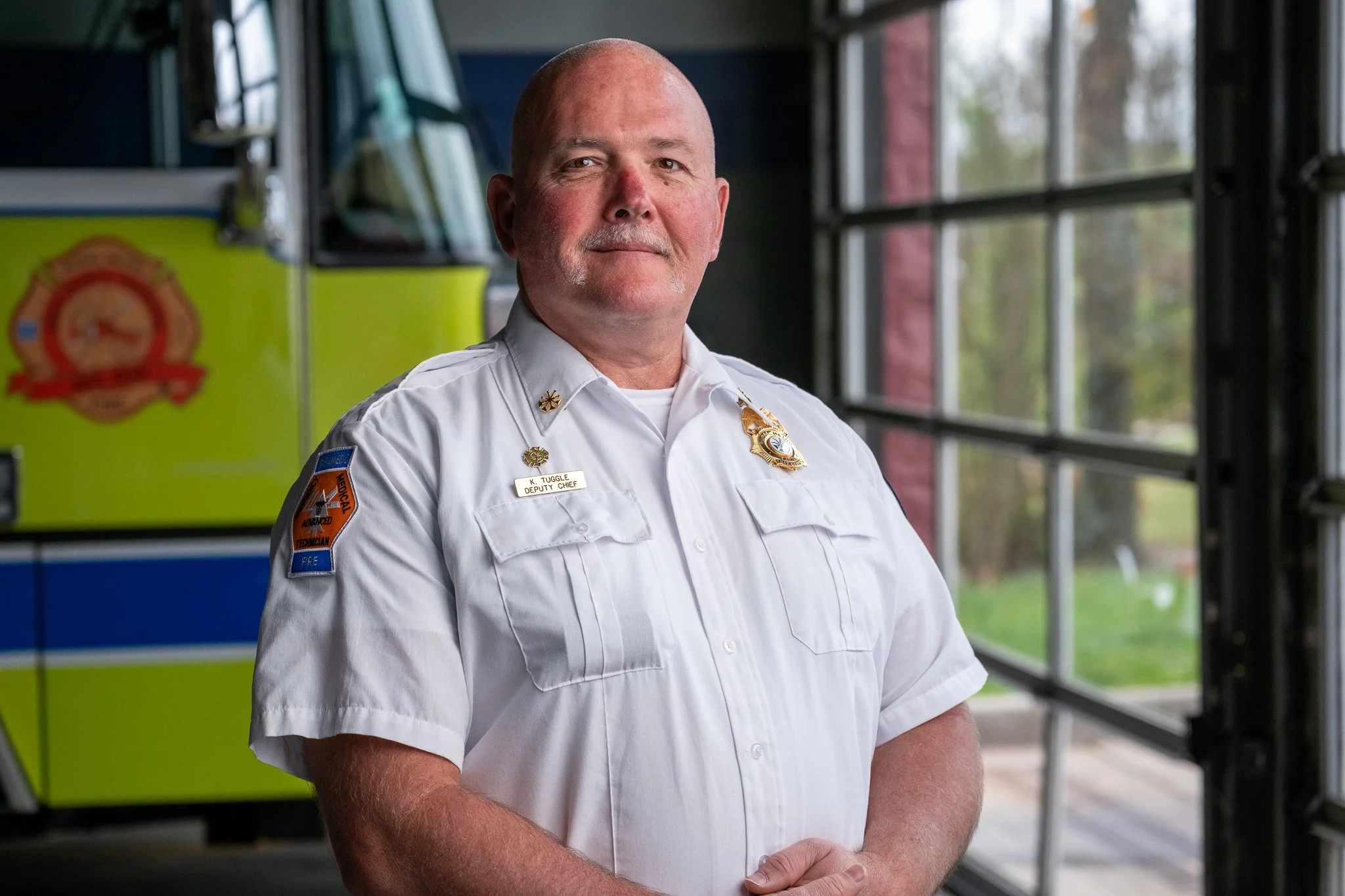 A middle-aged firefighter with a shaved head and a mustache, wearing a white uniform shirt with a badge and a name tag that reads "K. Tuggle Deputy Chief," standing indoors in front of a yellow emergency vehicle and large windows showing trees outside.