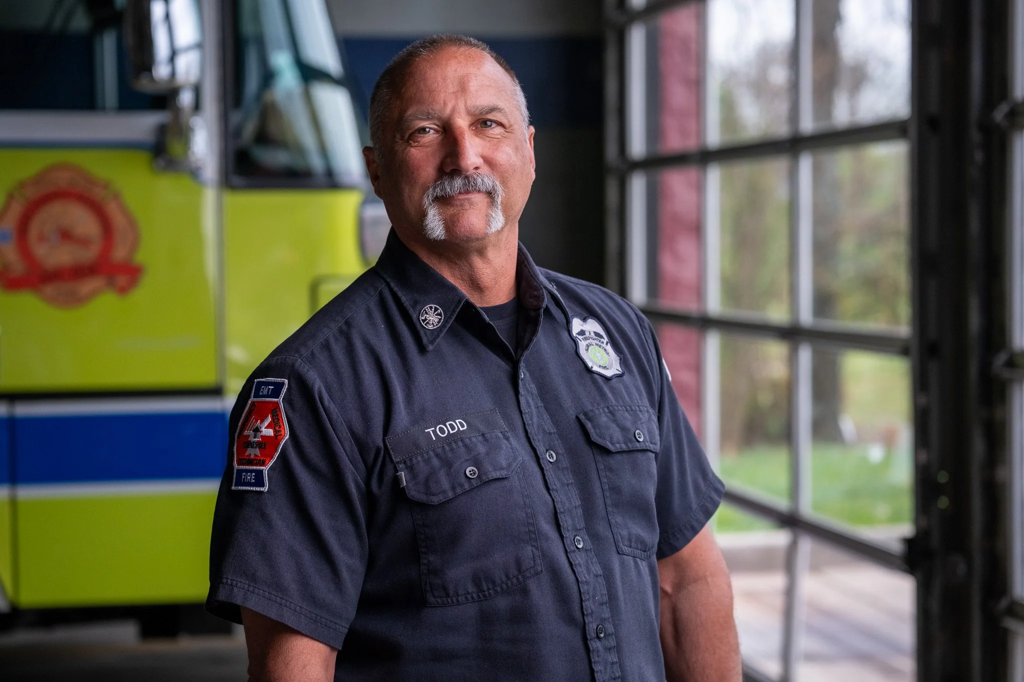 A middle-aged man with a grey mustache and goatee wearing a firefighter uniform standing inside a fire station.