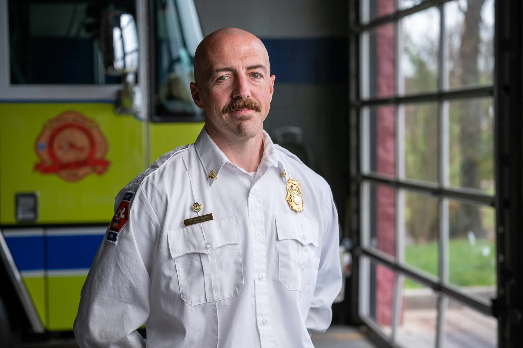 A firefighter standing in front of a fire truck and open garage doors, wearing a white uniform with badges and insignia.