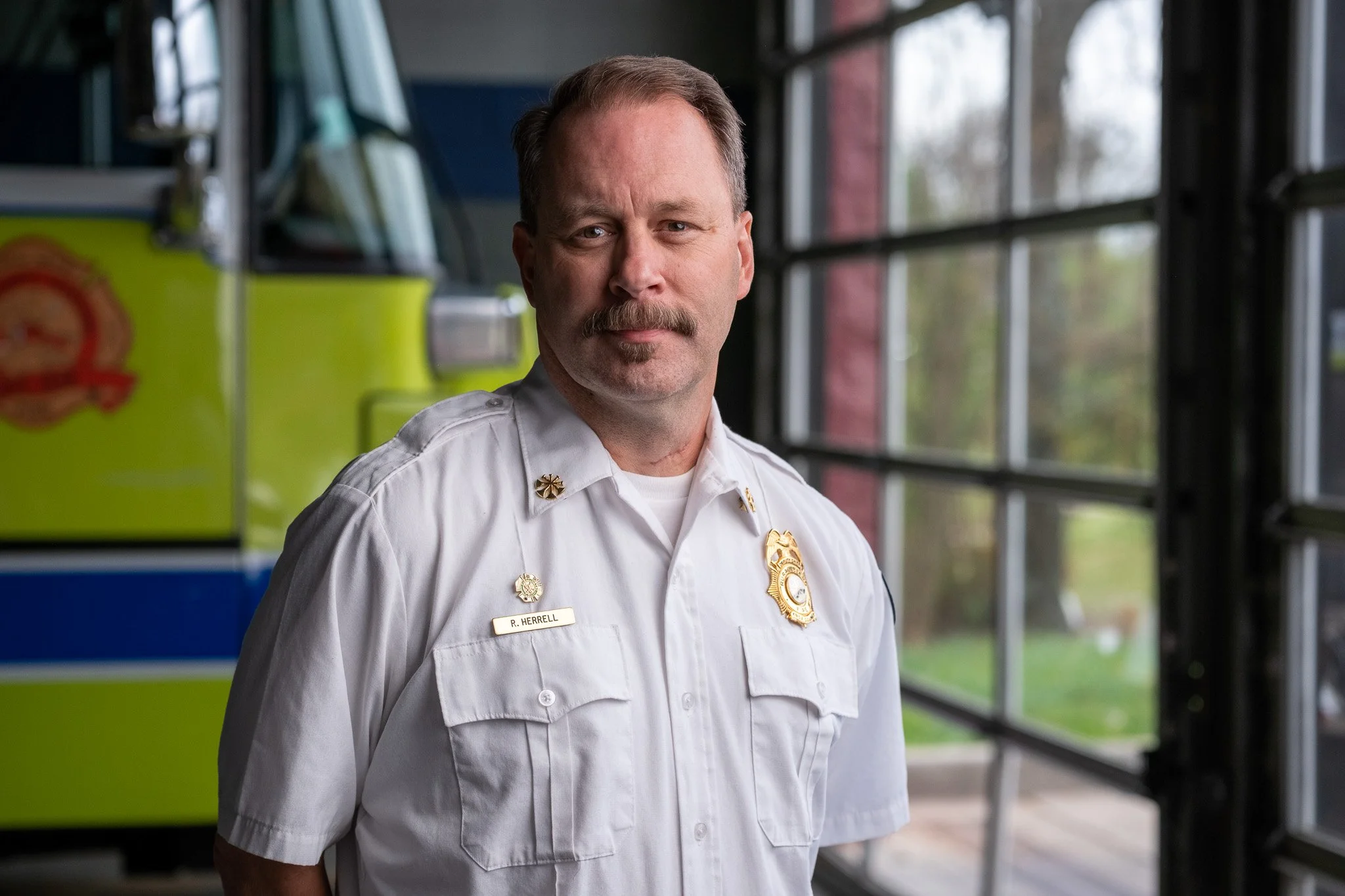 A firefighter in uniform standing inside a fire station near a fire truck. The firefighter is looking at the camera with a serious expression. The background shows large windows and part of the fire truck.