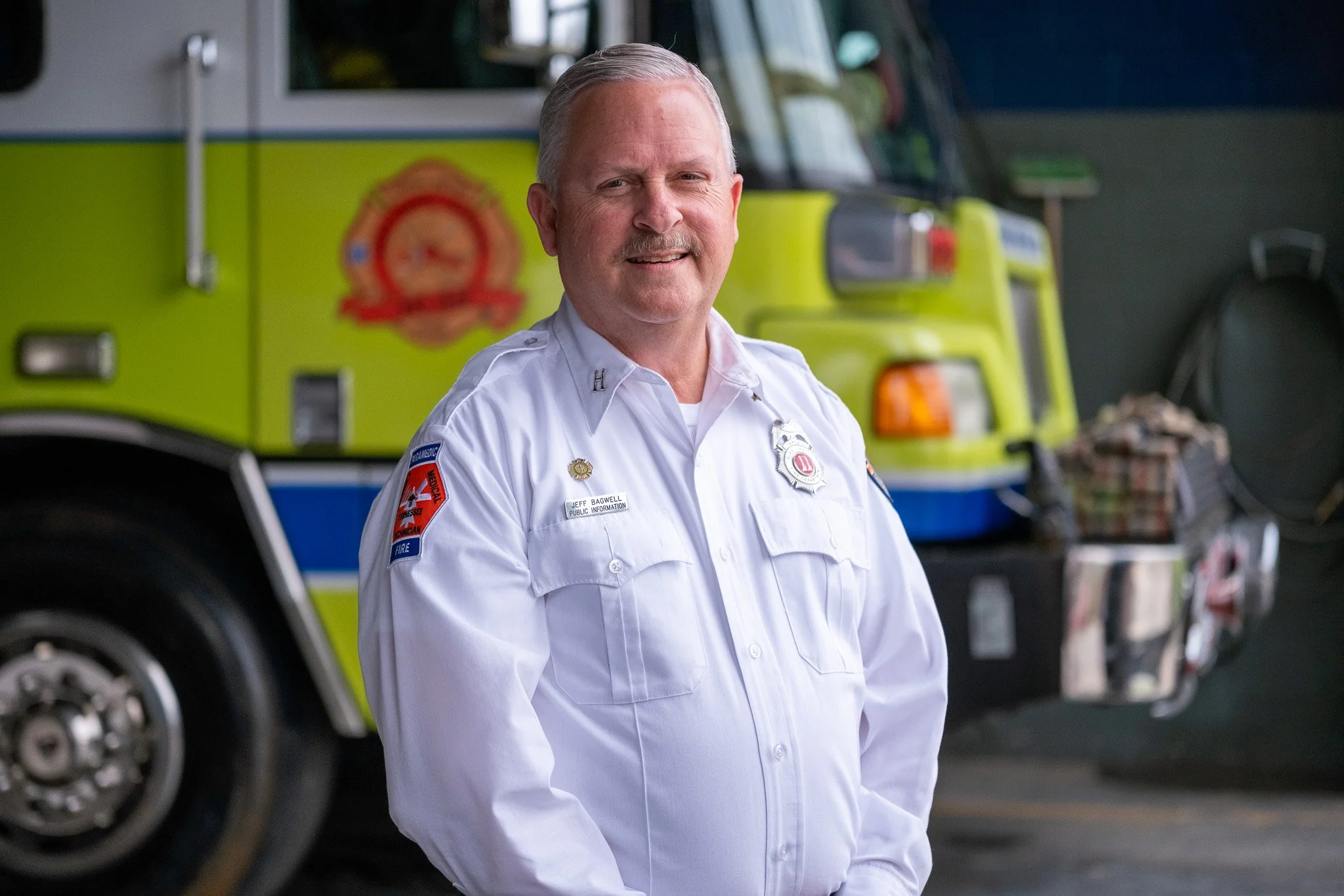 A smiling man in a white firefighter uniform standing in front of a yellow fire truck.
