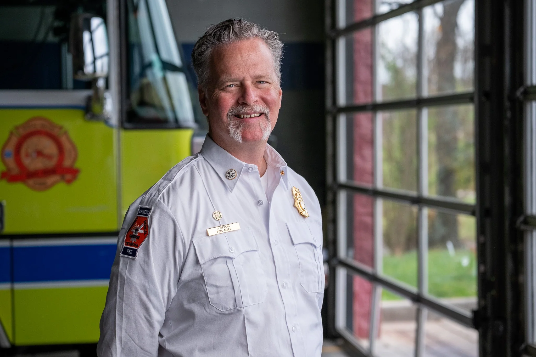 Smiling white male firefighter in uniform standing inside fire station with fire truck and large windows in the background.