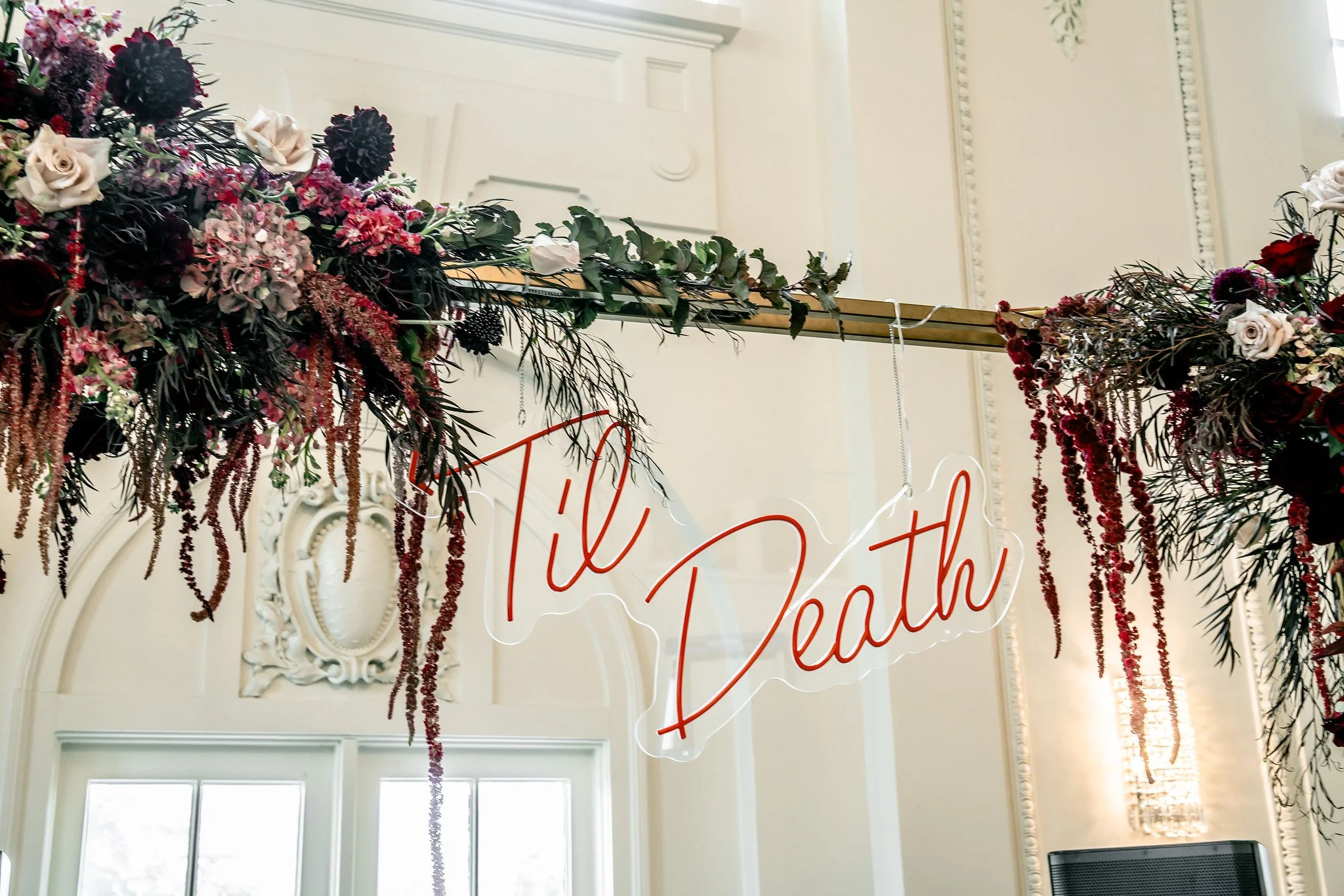 Floral arrangement with dark red, white, pink, and black flowers hanging from a horizontal bar. Neon sign with cursive text partially visible behind the flowers, in a room with ornate white walls and high windows.