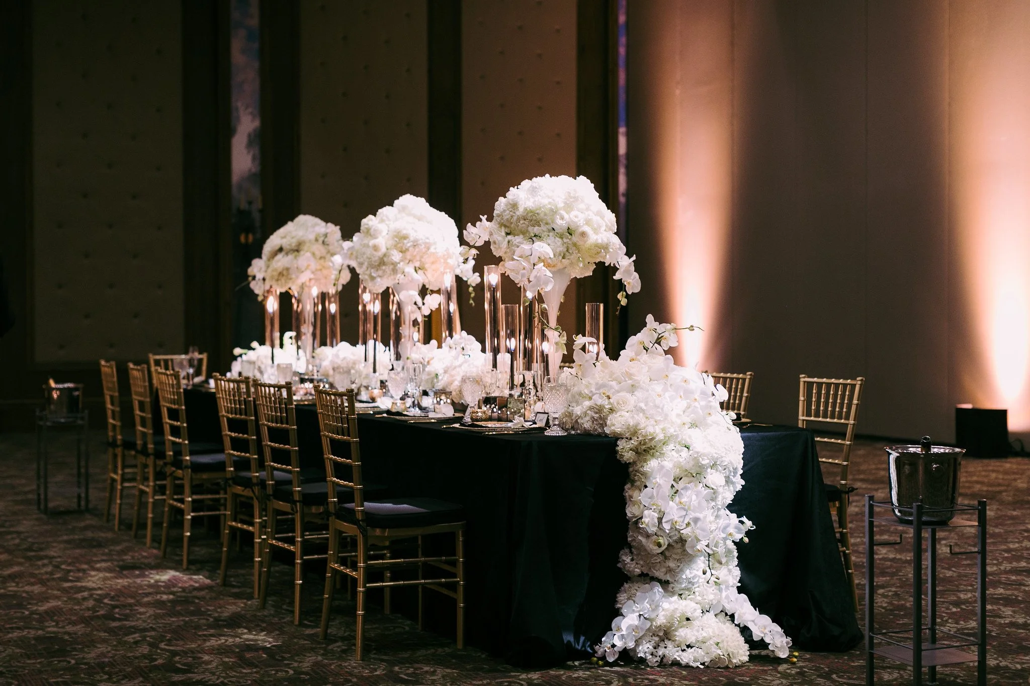 Elegant banquet table decorated with large white floral arrangements, glass candle holders, and gold chairs in a dimly lit room with warm lighting on the walls.