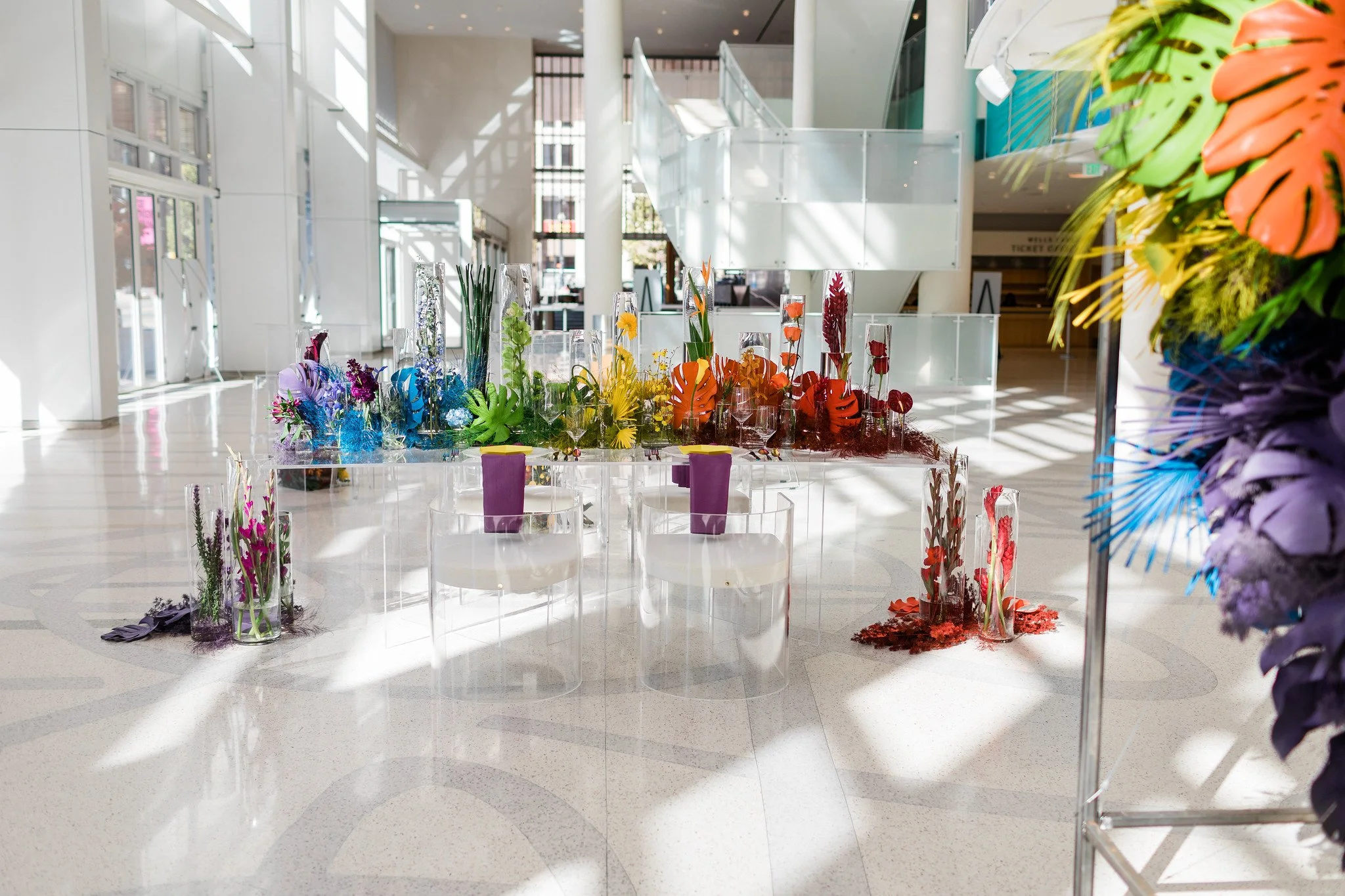An indoor space with a modern design, featuring a colorful floral art installation and glass display cases, illuminated by sunlight through large windows.