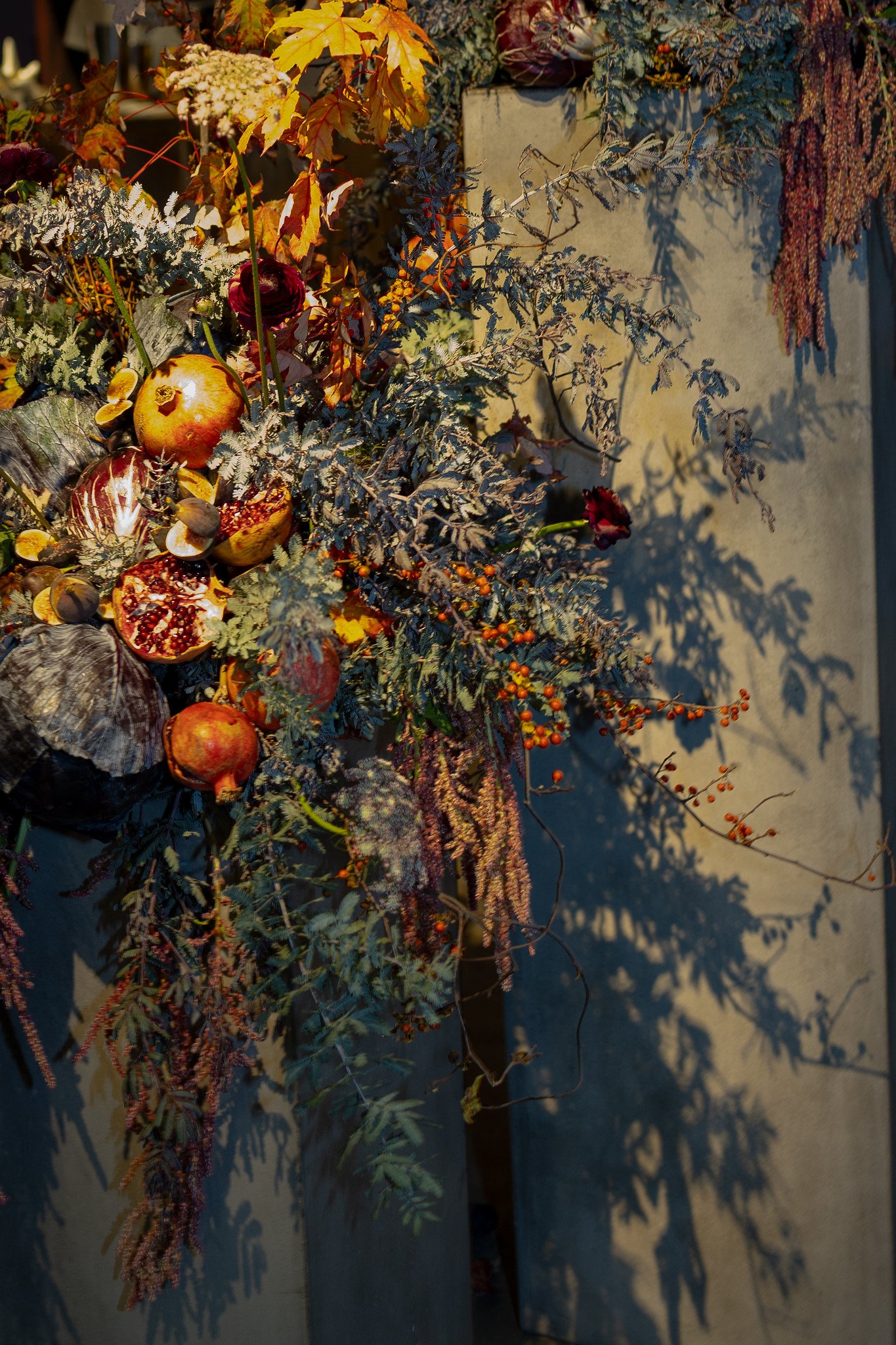 Autumn-themed floral arrangement with pomegranates, dried leaves, and various flowers, casting shadows on a gray surface.
