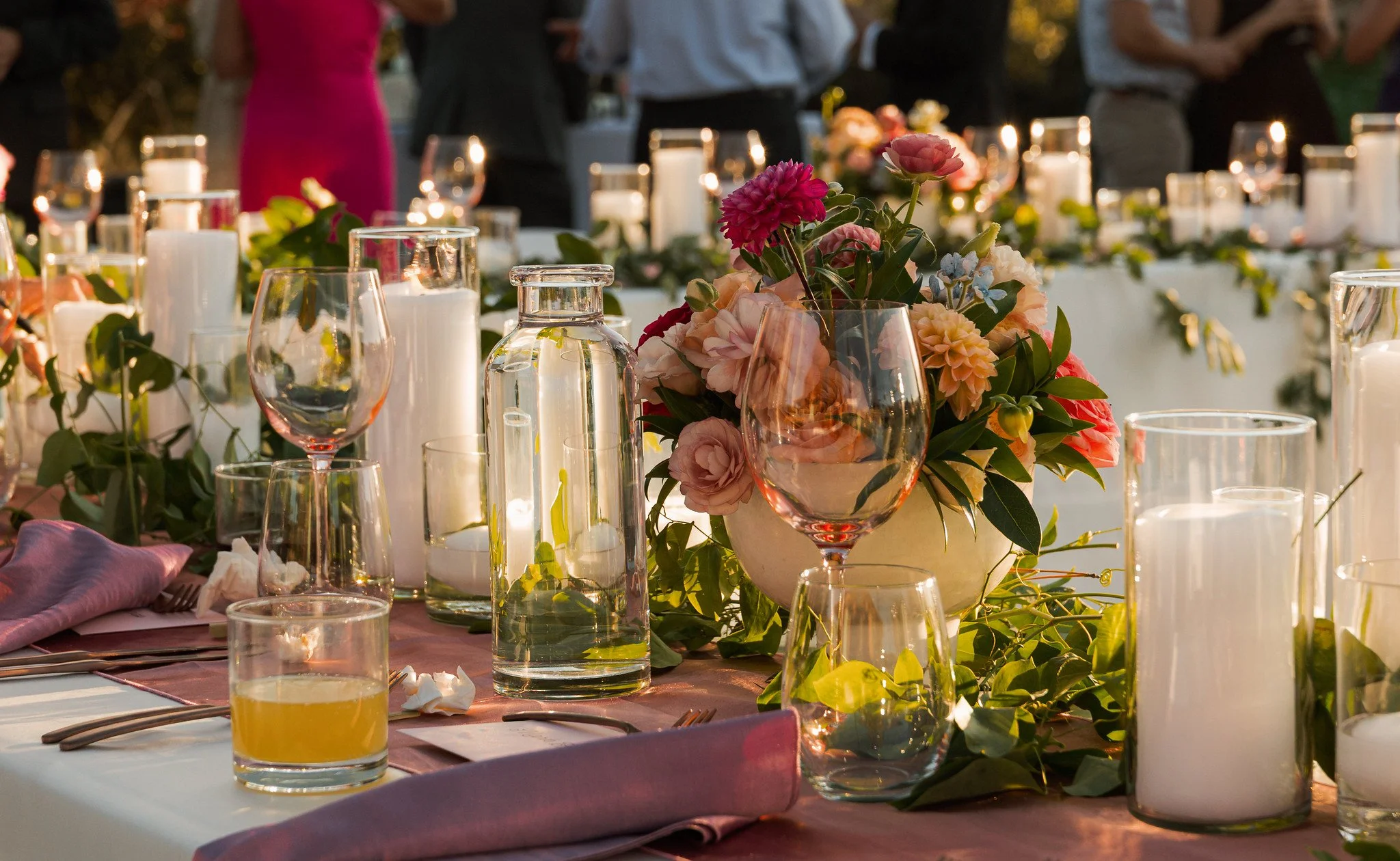 A decorated outdoor banquet table with floral centerpieces, candles, and glassware, set for an evening event.