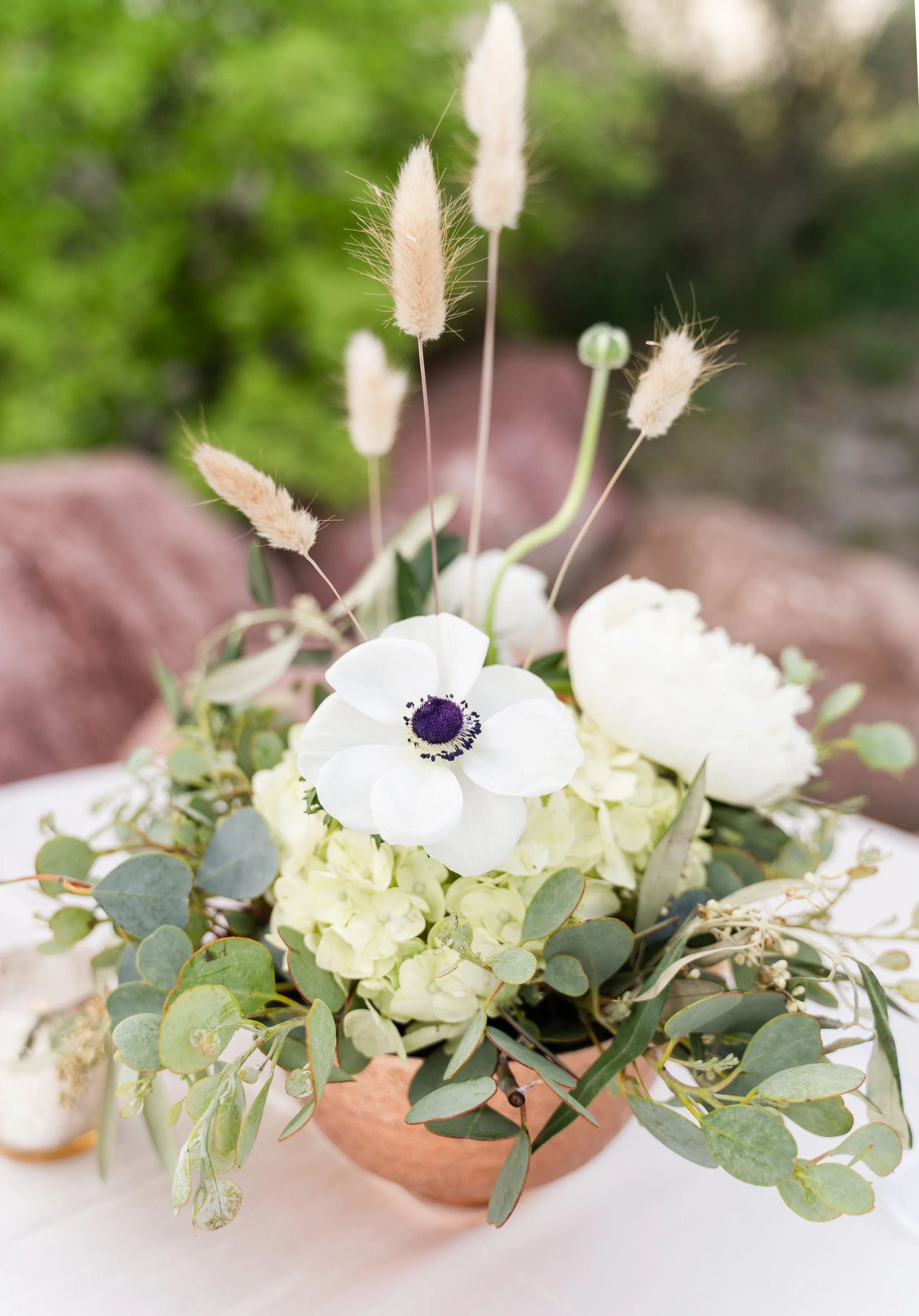 A floral arrangement featuring white flowers, green hydrangeas, and eucalyptus leaves in a terracotta pot, set against a blurred outdoor background.