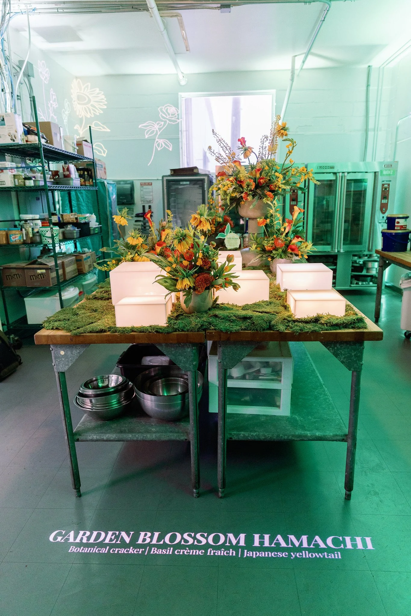 Table decorated with floral arrangements, illuminated white blocks, and moss, inside a kitchen or bakery with shelves of supplies and green industrial refrigerators. The floor has a sign with pink text that reads, 'GARDEN BLOSSOM HAMACHI Botanical cr